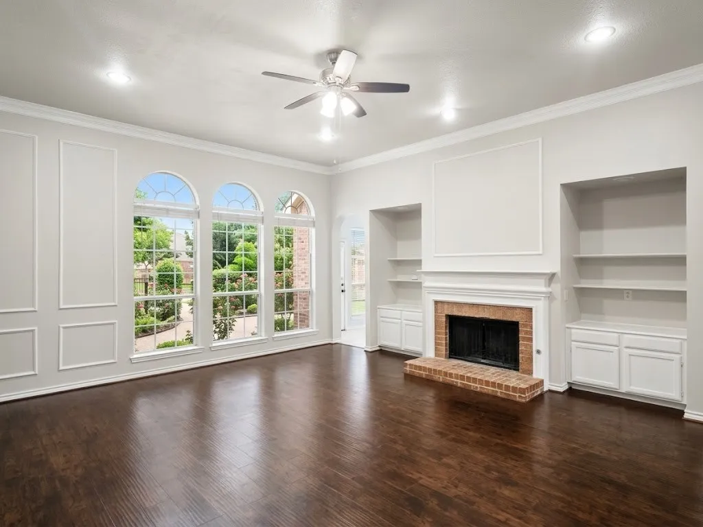 Hardwood flooring, three arched windows, a brick fireplace, built-in shelving, and a ceiling fan