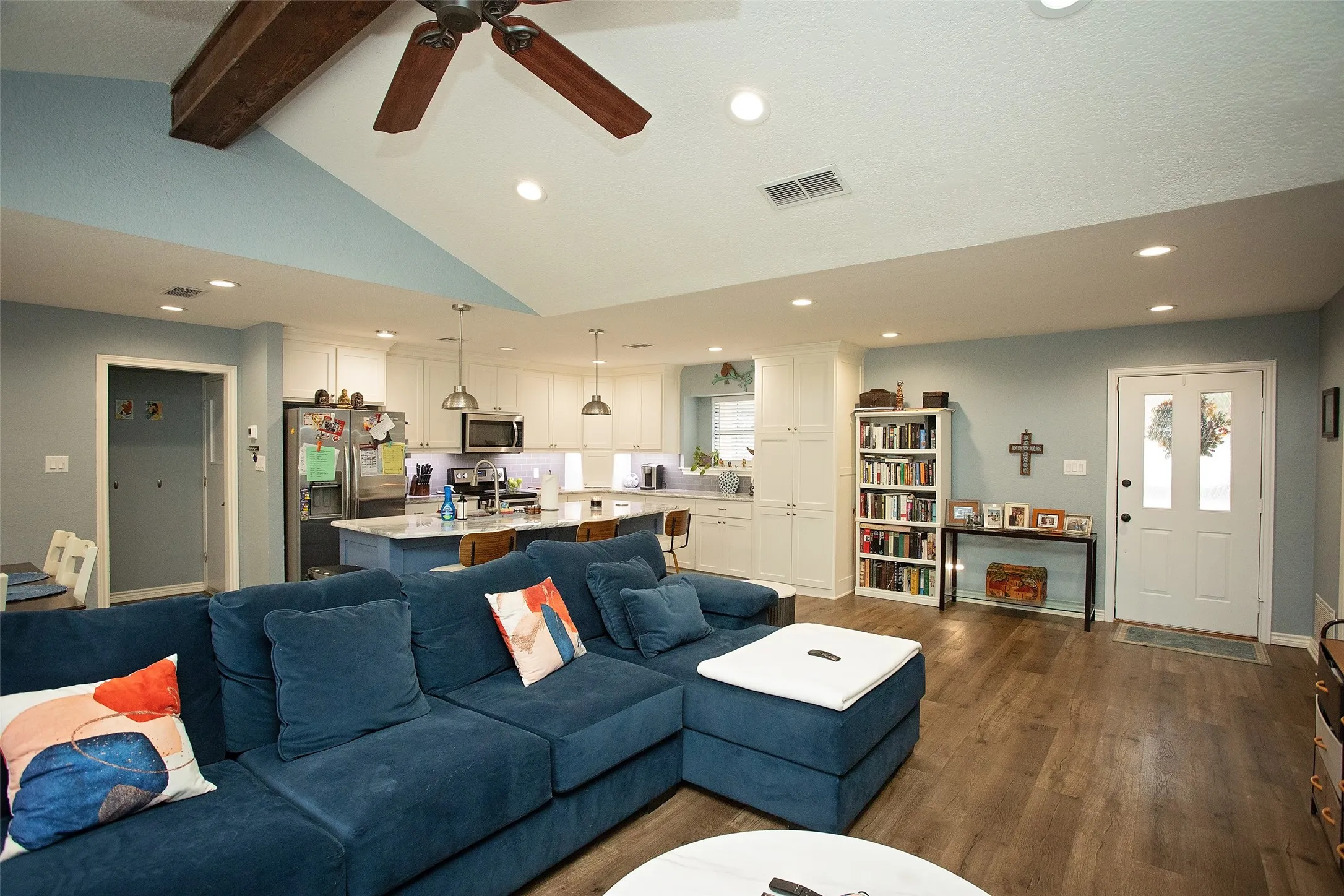 Vaulted ceiling with exposed wood beam and ceiling fan, recessed lighting, wood-finish flooring, and a kitchen featuring white cabinetry and a central island with pendant lighting