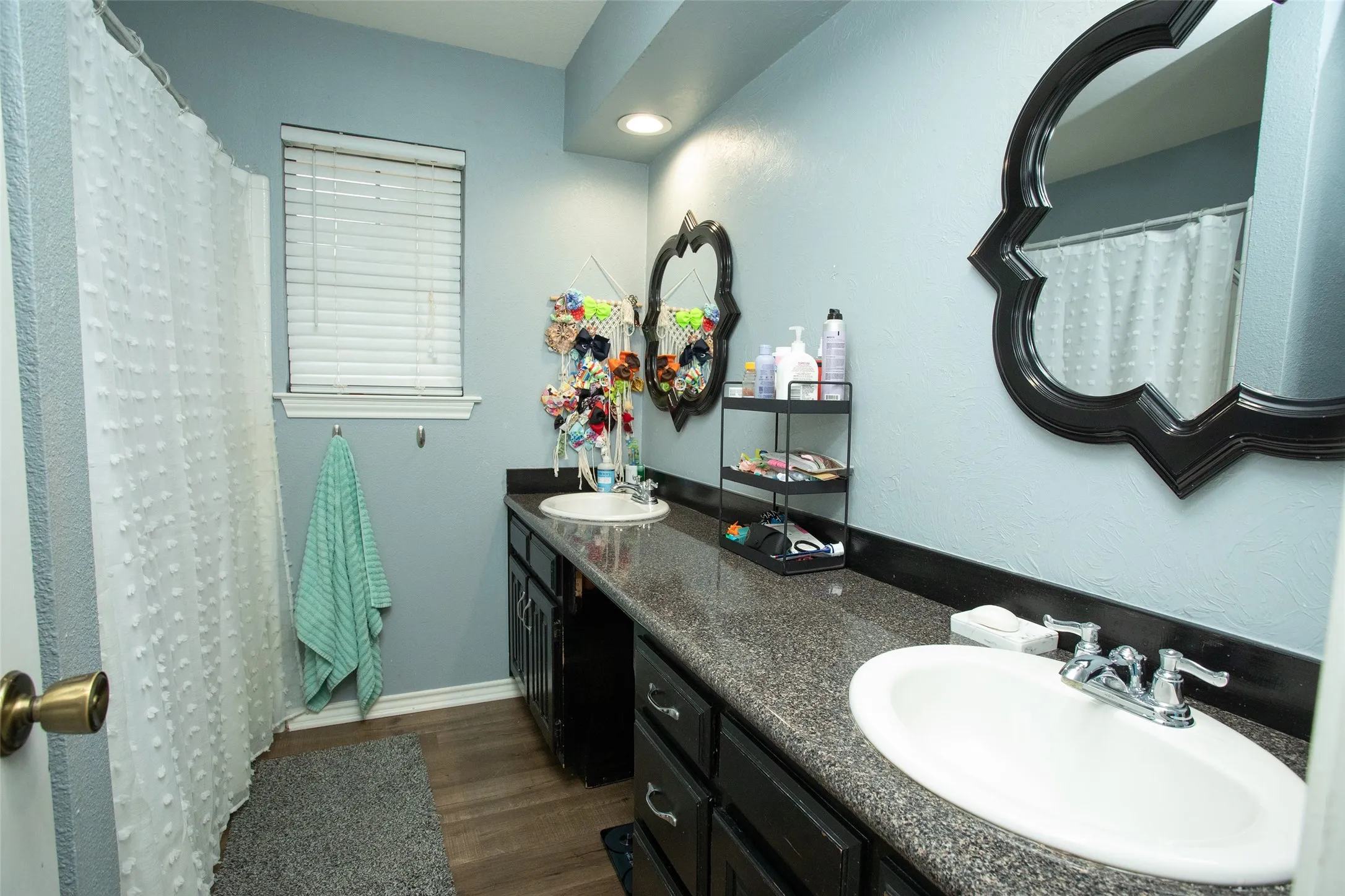 Dual vanity with undermount sinks, speckled countertop, dark wood-finish cabinetry, wood-finish flooring, and a window with blinds