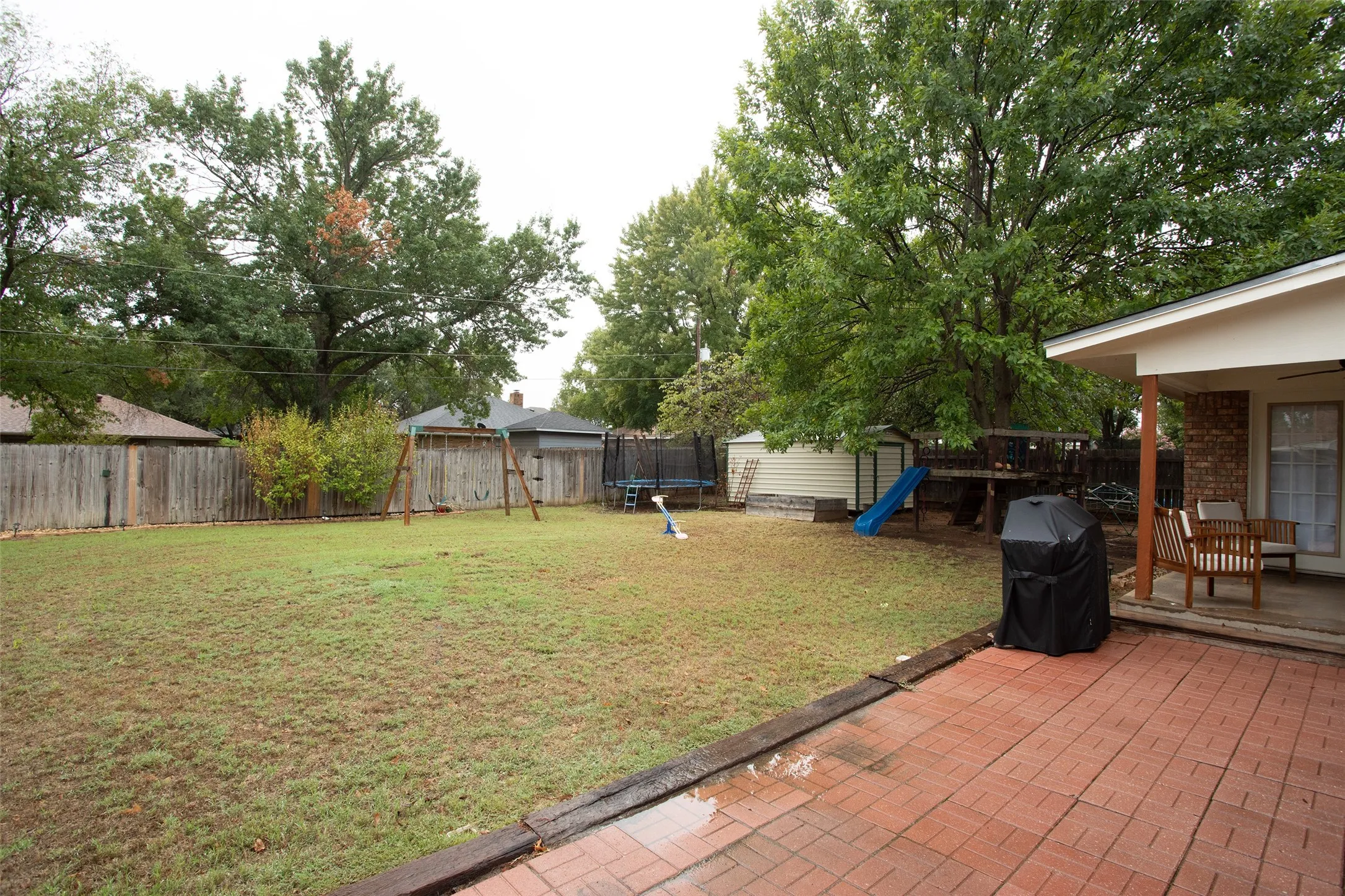 Expansive backyard featuring a red brick patio, covered outdoor living space, and mature trees