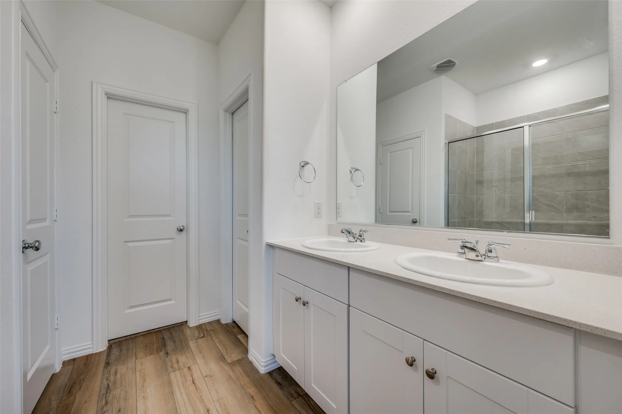 Double vanity with undermount sinks, light-toned countertop, wood-finish flooring, and a glass-enclosed shower with tiled walls