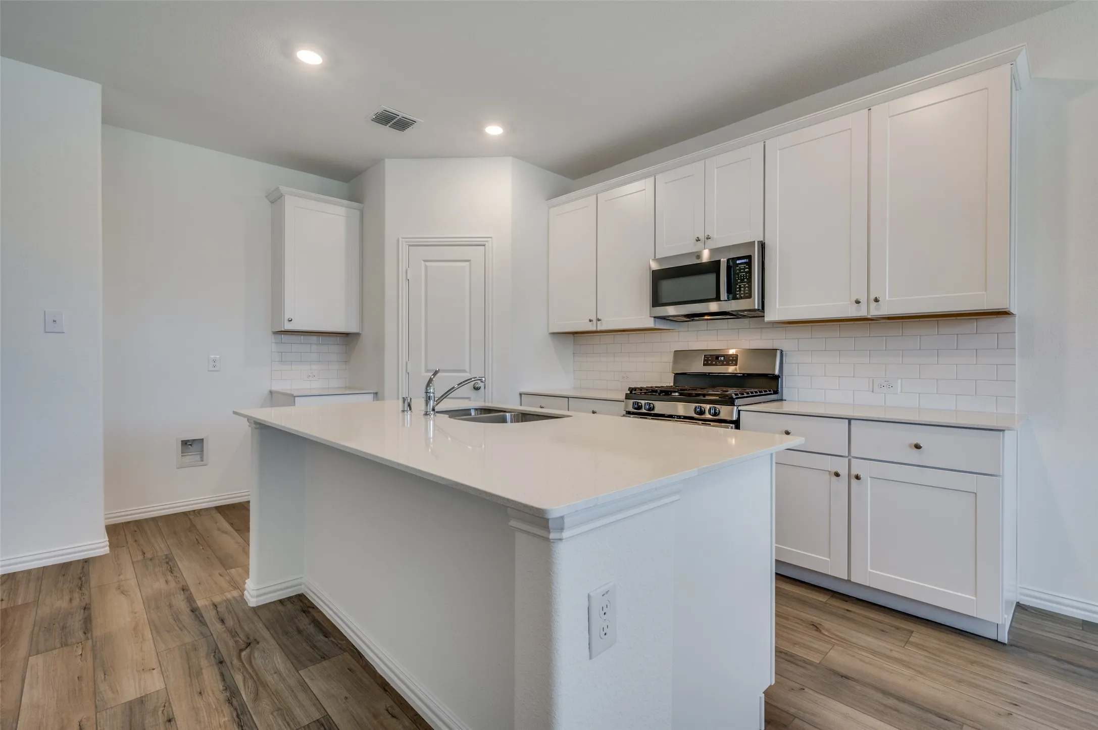 Kitchen featuring a center island with double basin sink, stainless steel gas range, white shaker cabinetry, subway tile backsplash, and wood-finish flooring