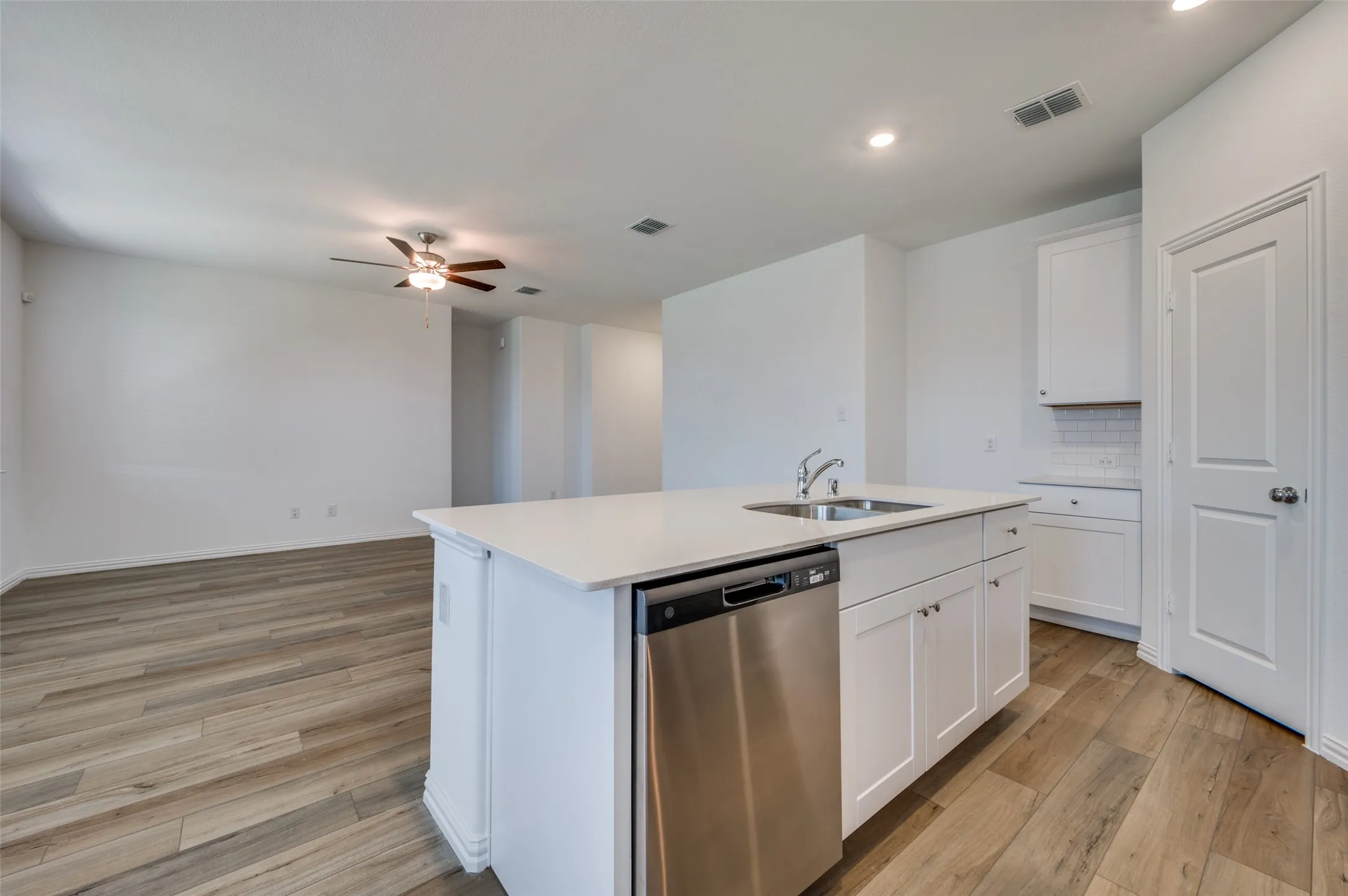 Open-concept kitchen featuring a center island with sink, white shaker cabinetry, stainless steel dishwasher, and wood-finish flooring