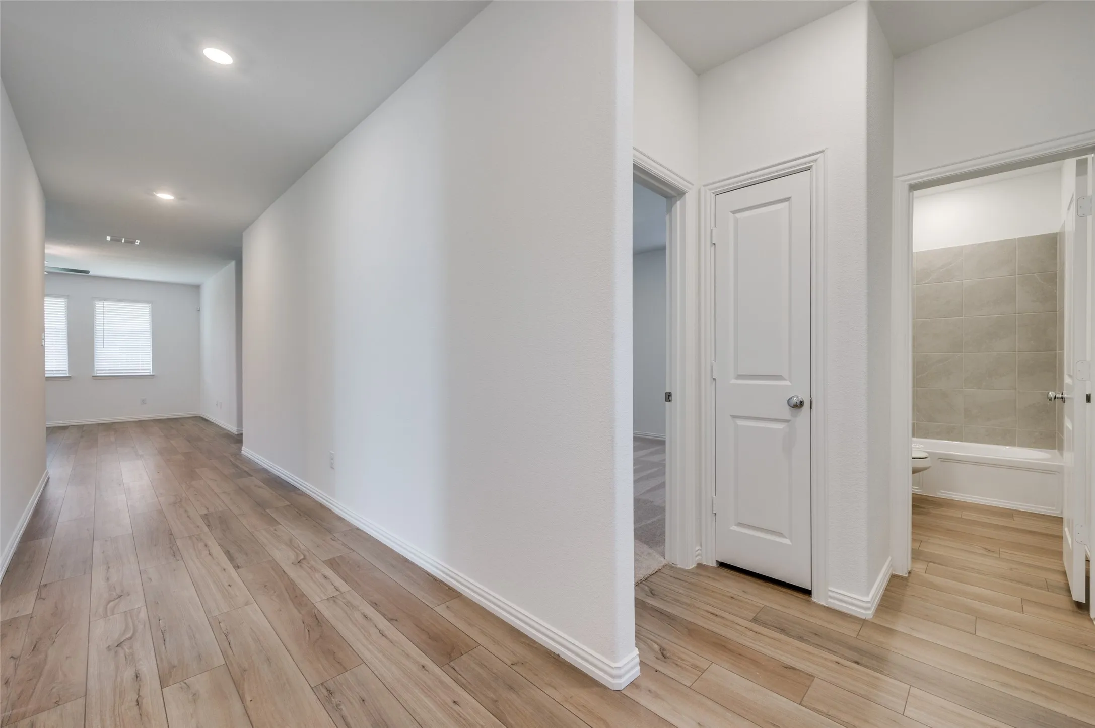 Wood-finish flooring throughout, white walls, recessed ceiling lighting, and a bathroom with a tiled tub surround