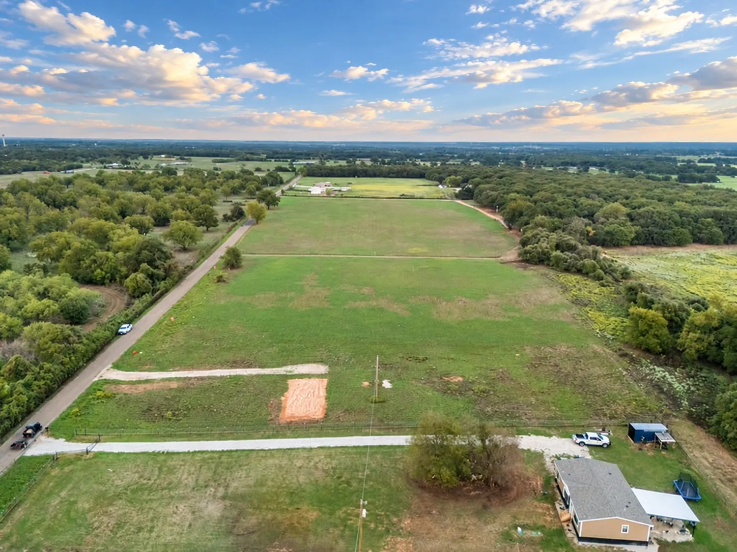 Expansive green fields bordered by mature trees and a paved road, featuring a single-story residence with a covered porch
