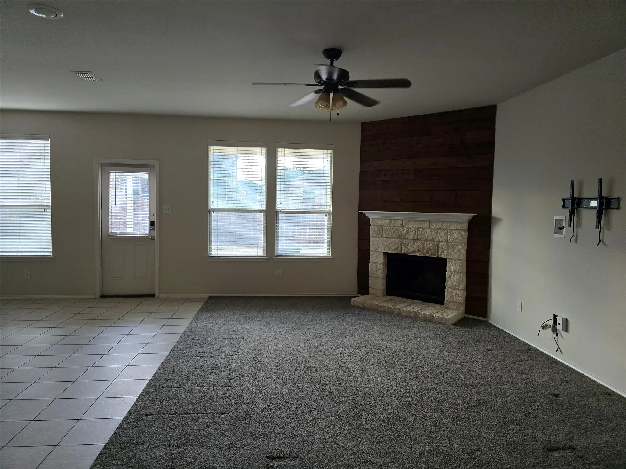 Built-in fireplace with stone surround, wood-paneled accent wall, dual windows with blinds, and a ceiling fan with integrated lighting