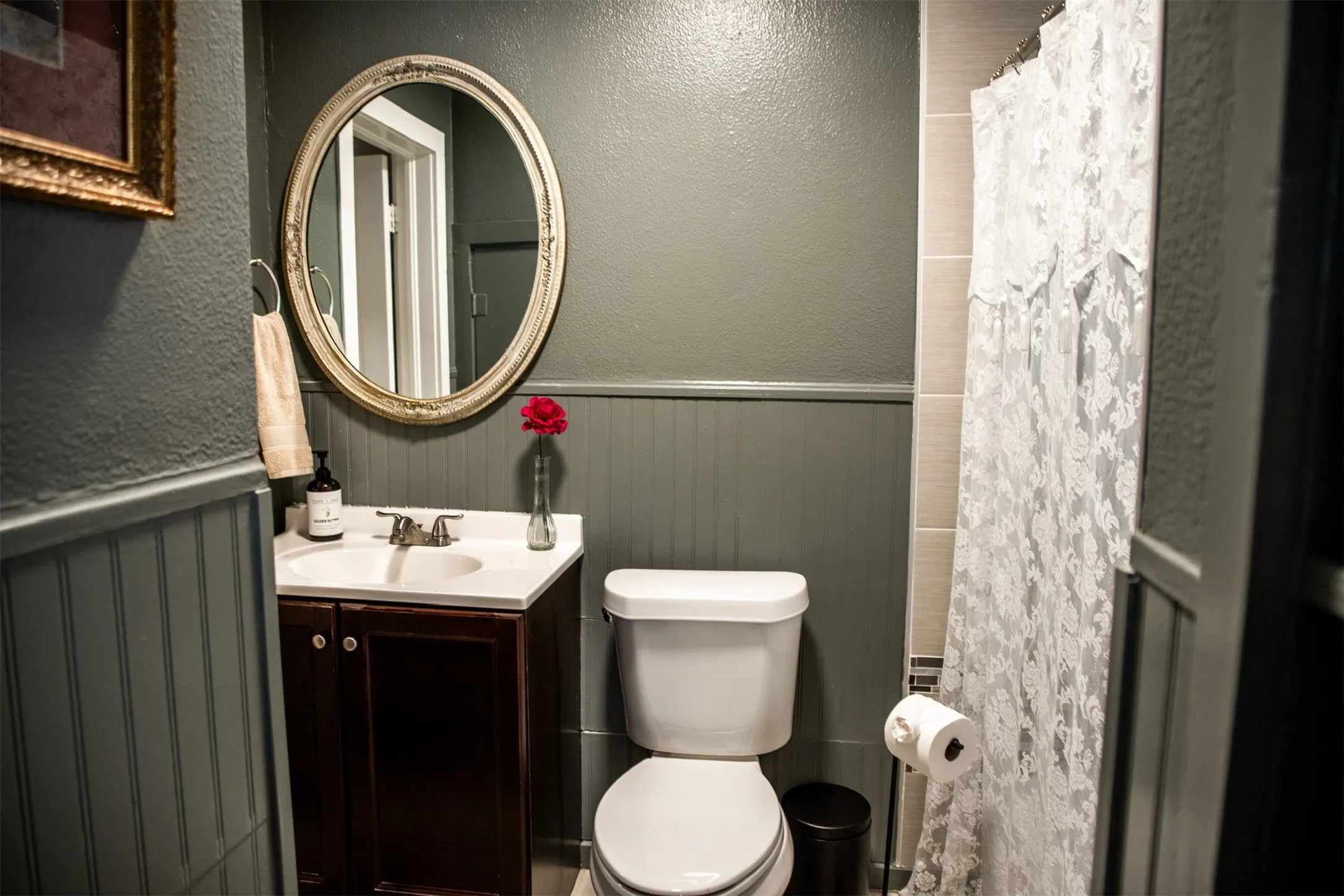 Beadboard wainscoting, oval-framed mirror, vanity with integrated sink, toilet, and tiled shower area