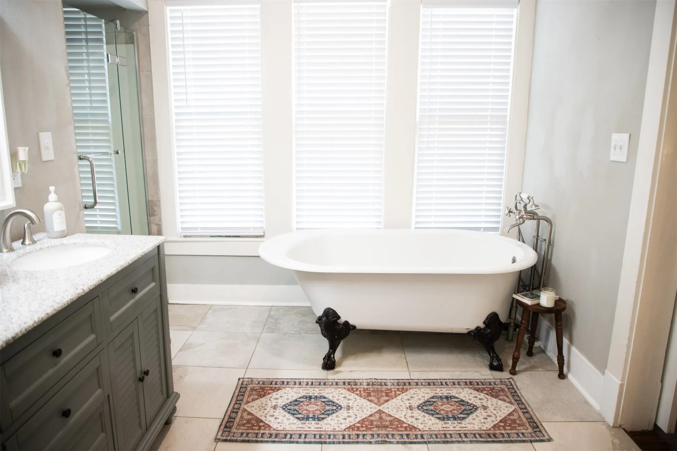 Clawfoot soaking tub, single vanity with integrated sink, glass-enclosed shower, three windows with blinds, and tile flooring