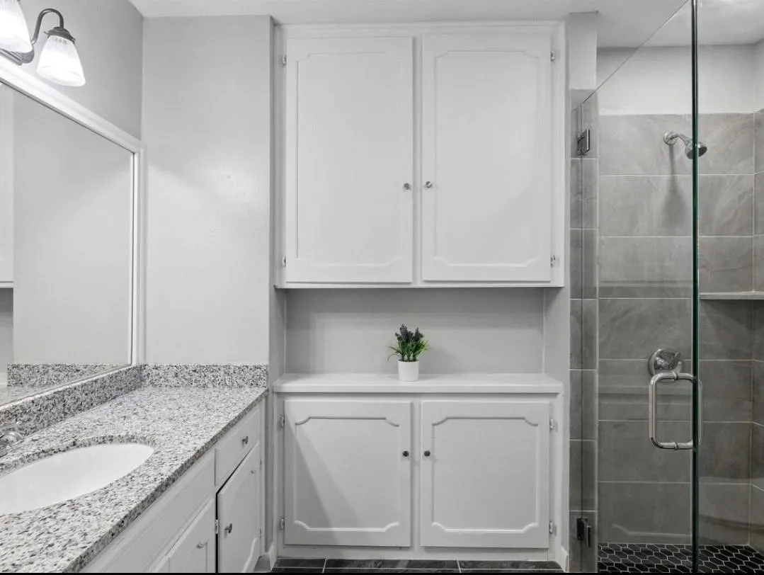 Built-in cabinetry with white shaker-style doors, granite-style countertop, integrated sink, mirrored vanity, and a glass-enclosed shower with tiled walls and a hex tile floor