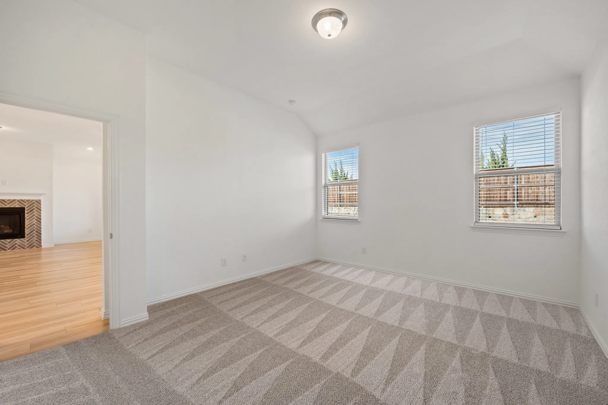 Vaulted ceiling with flush-mount lighting, patterned carpet flooring, two windows with blinds, and white trim throughout