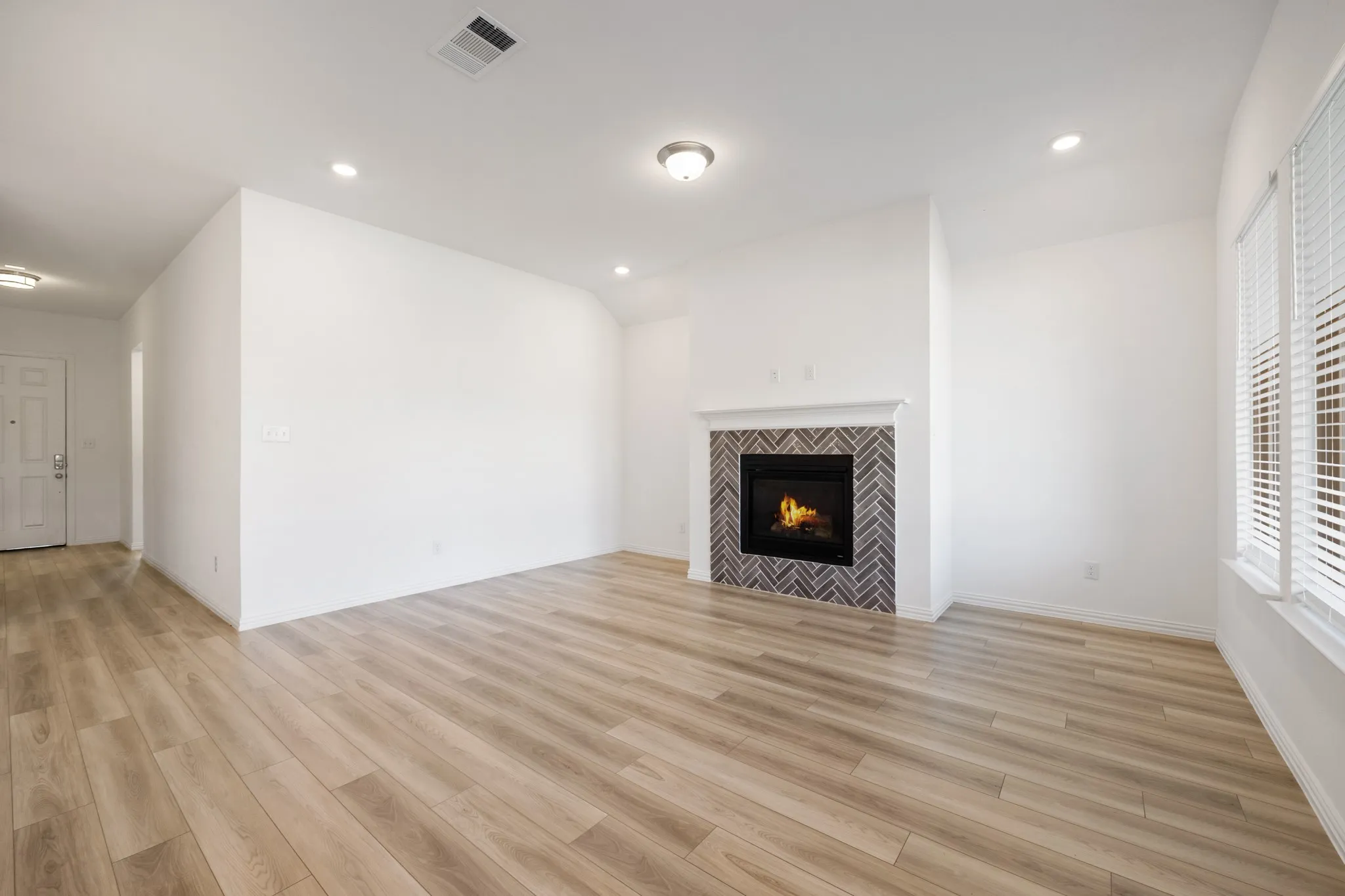 Spacious living area featuring wood-finish flooring, a fireplace with a herringbone tile surround, multiple recessed light fixtures, and windows with blinds