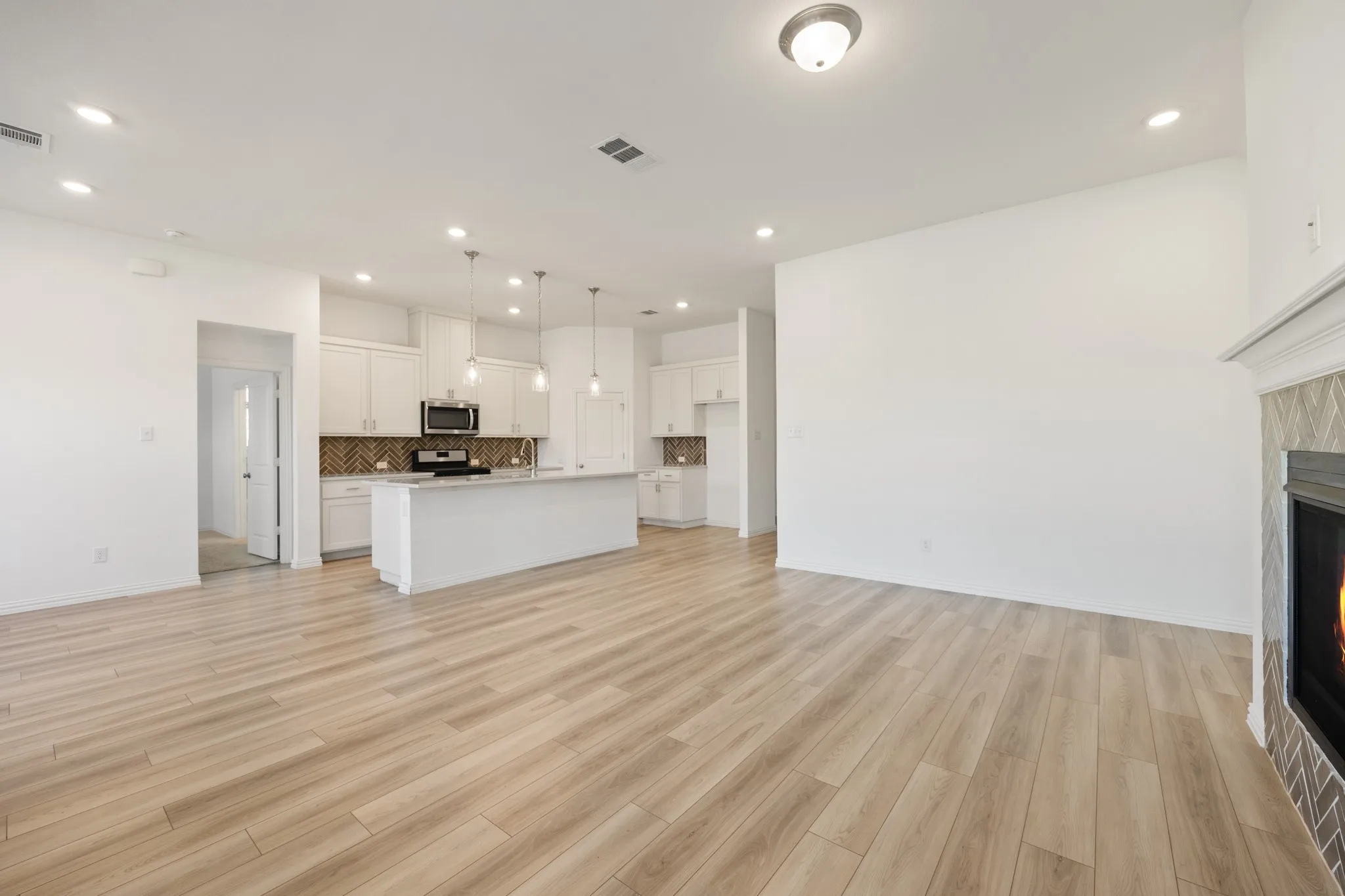 Open-concept living space featuring wood-finish flooring, a kitchen island with pendant lighting, white cabinetry, a herringbone tile backsplash, and a fireplace with a decorative mantel and tile surround