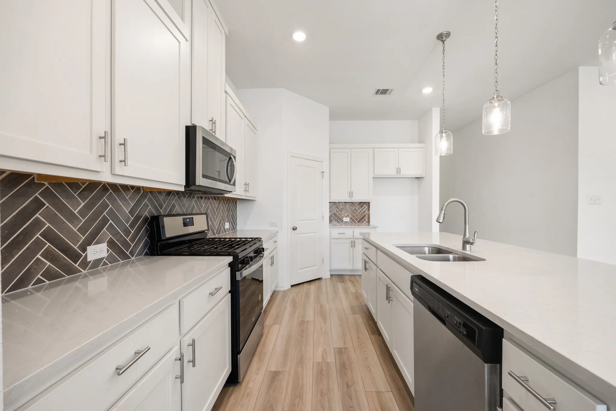 White shaker-style cabinetry with brushed nickel hardware, stainless steel appliances, a dark herringbone tile backsplash, and light wood-finish flooring