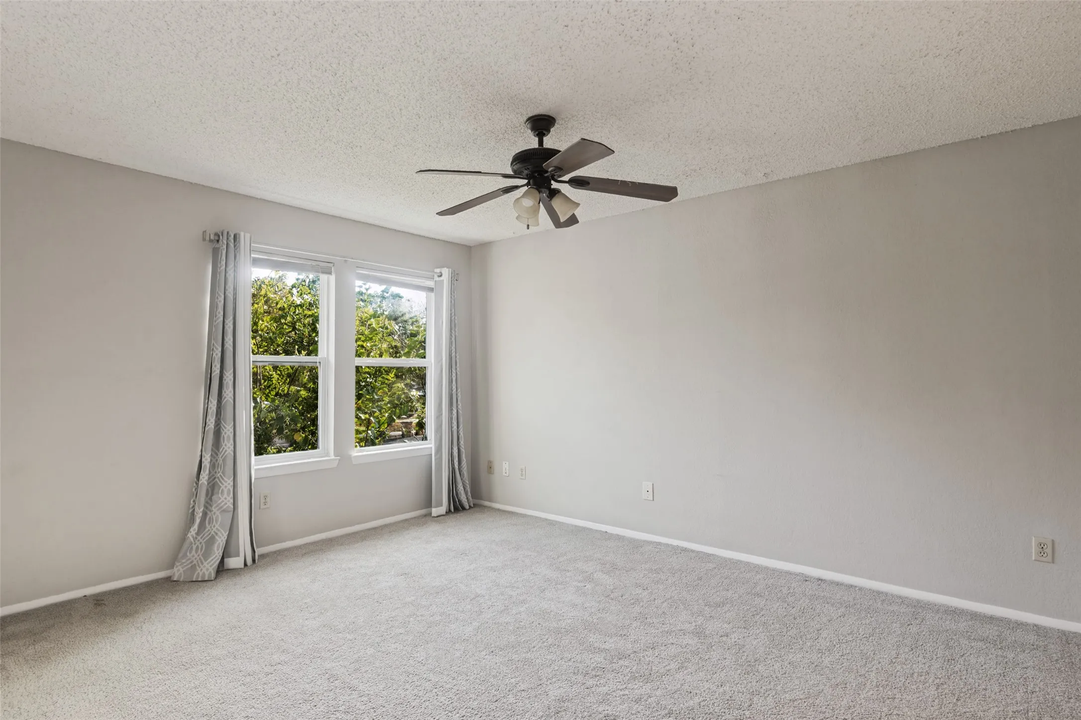 Plush gray carpeting throughout, featuring two windows with white framing and a ceiling fan with integrated​​‌​​​​‌​​‌‌​‌‌​​​‌‌​‌​‌​‌​​​‌​​ lighting