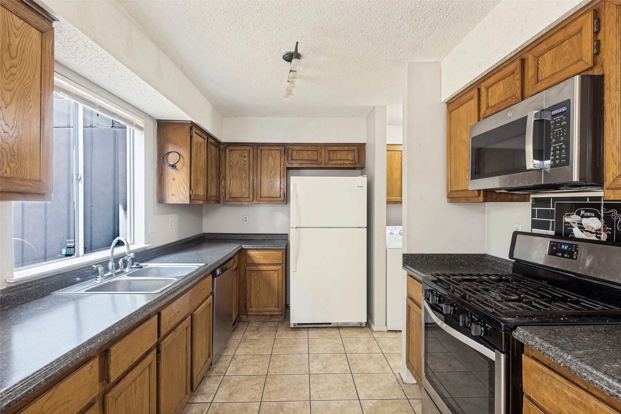 Kitchen featuring wood cabinetry, stainless steel microwave, gas range, double basin stainless steel sink, and tile flooring