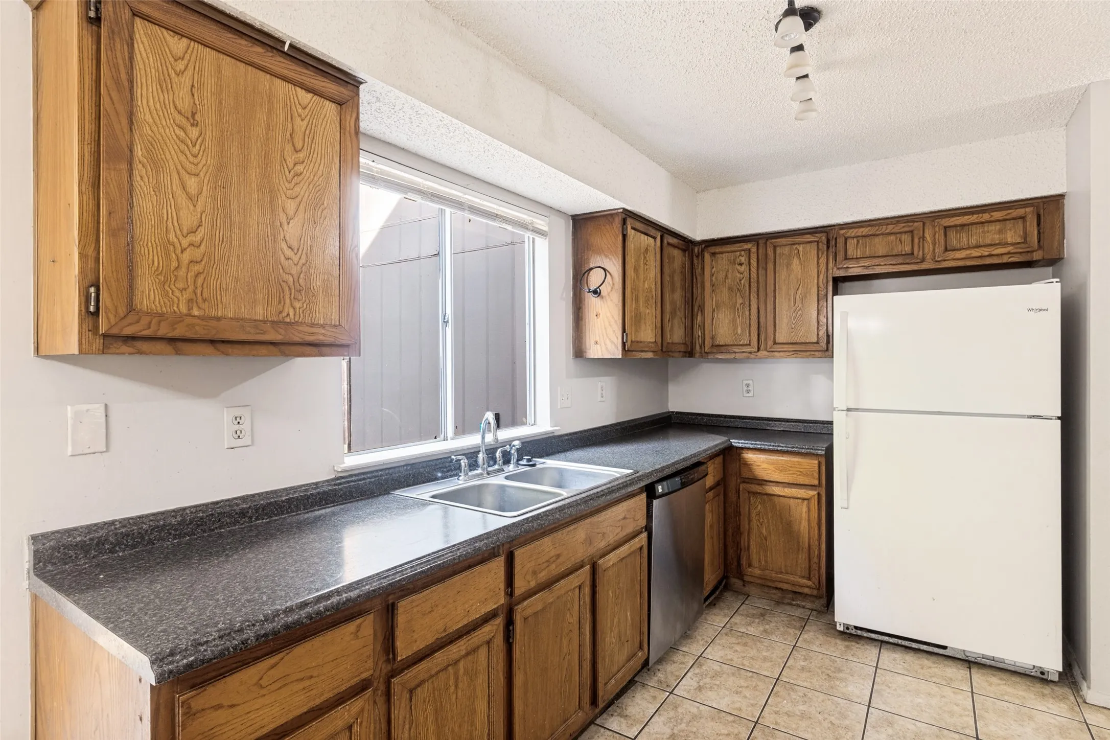 Galley kitchen featuring wood-finish cabinetry, dark stone countertops, stainless steel appliances including a gas range, and a white refrigerator