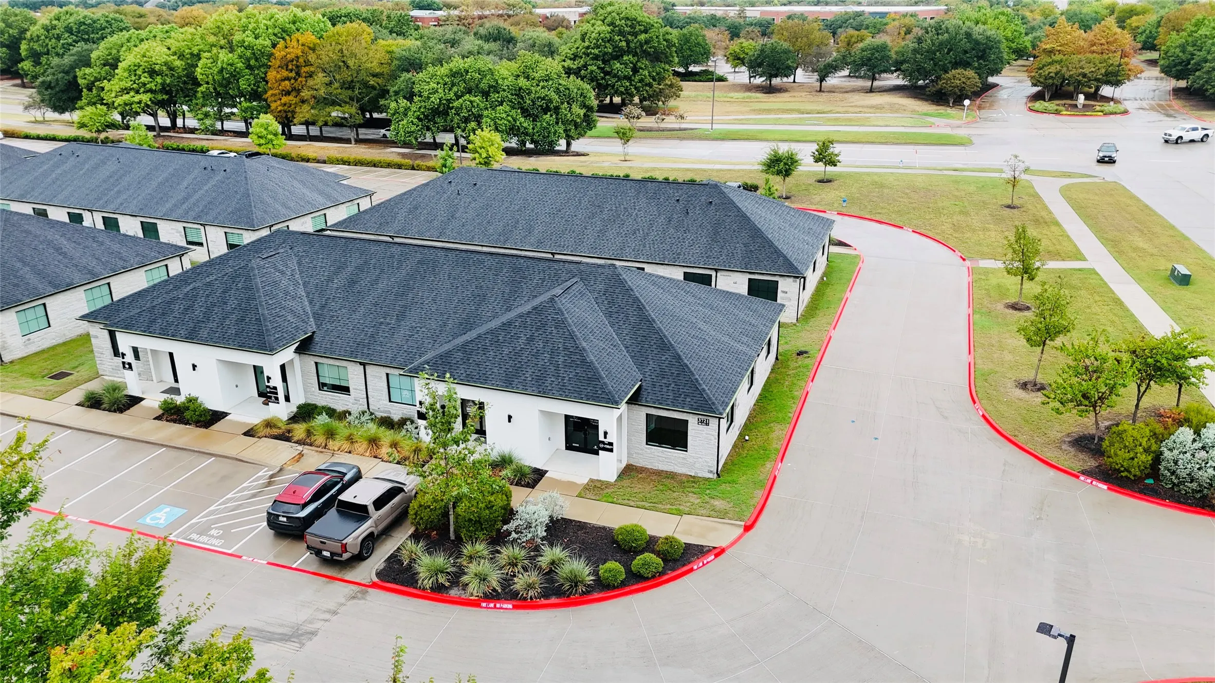 White stone exterior, dark shingled roofs, ample surface parking, paved driveways, and landscaped grounds