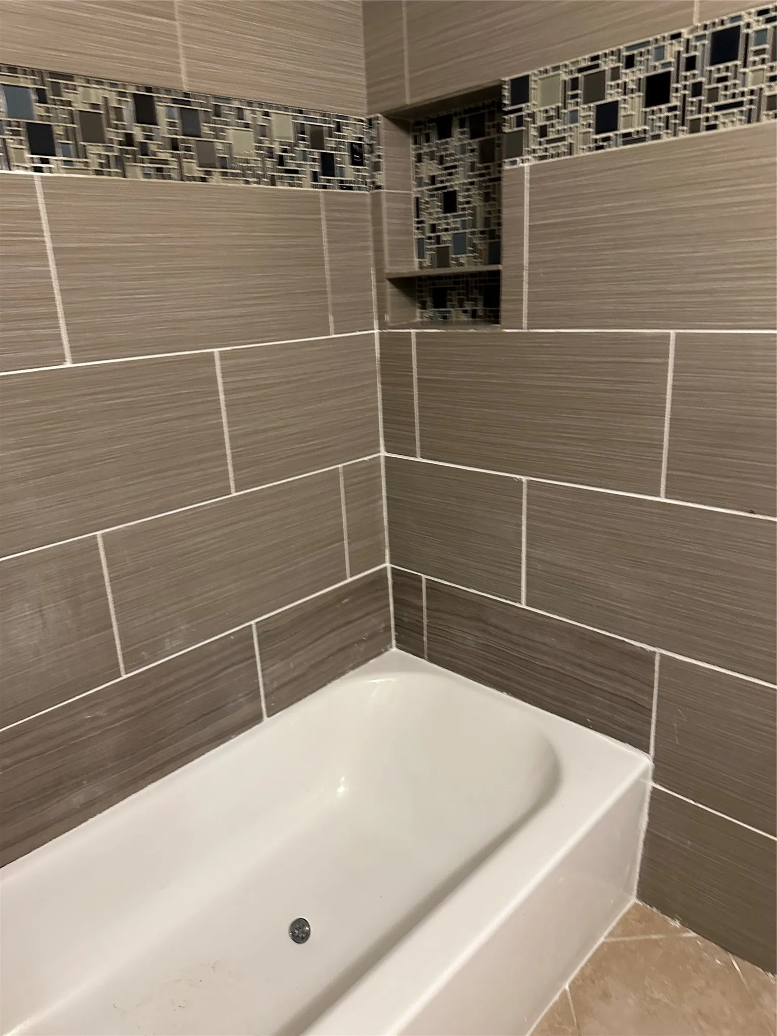 Bathtub with a mosaic tile accent band, built-in shelving, and large format wall tiles