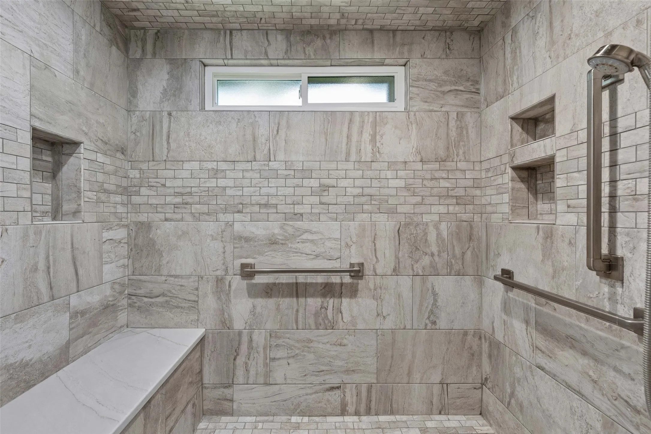 Oversized Walk-In Shower — The remodeled primary shower features expansive tilework, built-in niches, a bench, handheld shower fixture, and grab bars for added convenience and flexibility.