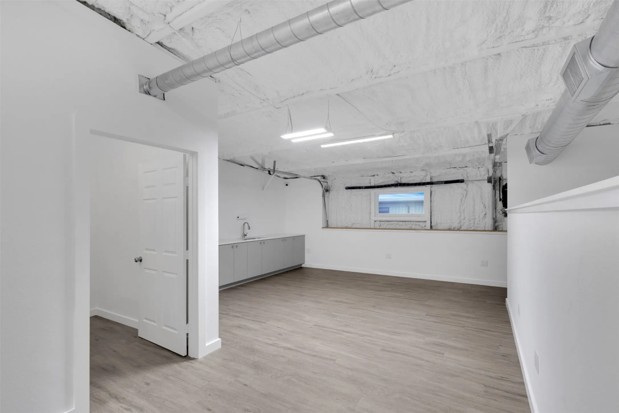 Spacious room featuring wood-finish flooring, white walls, built-in cabinetry with a sink, and exposed ceiling ductwork