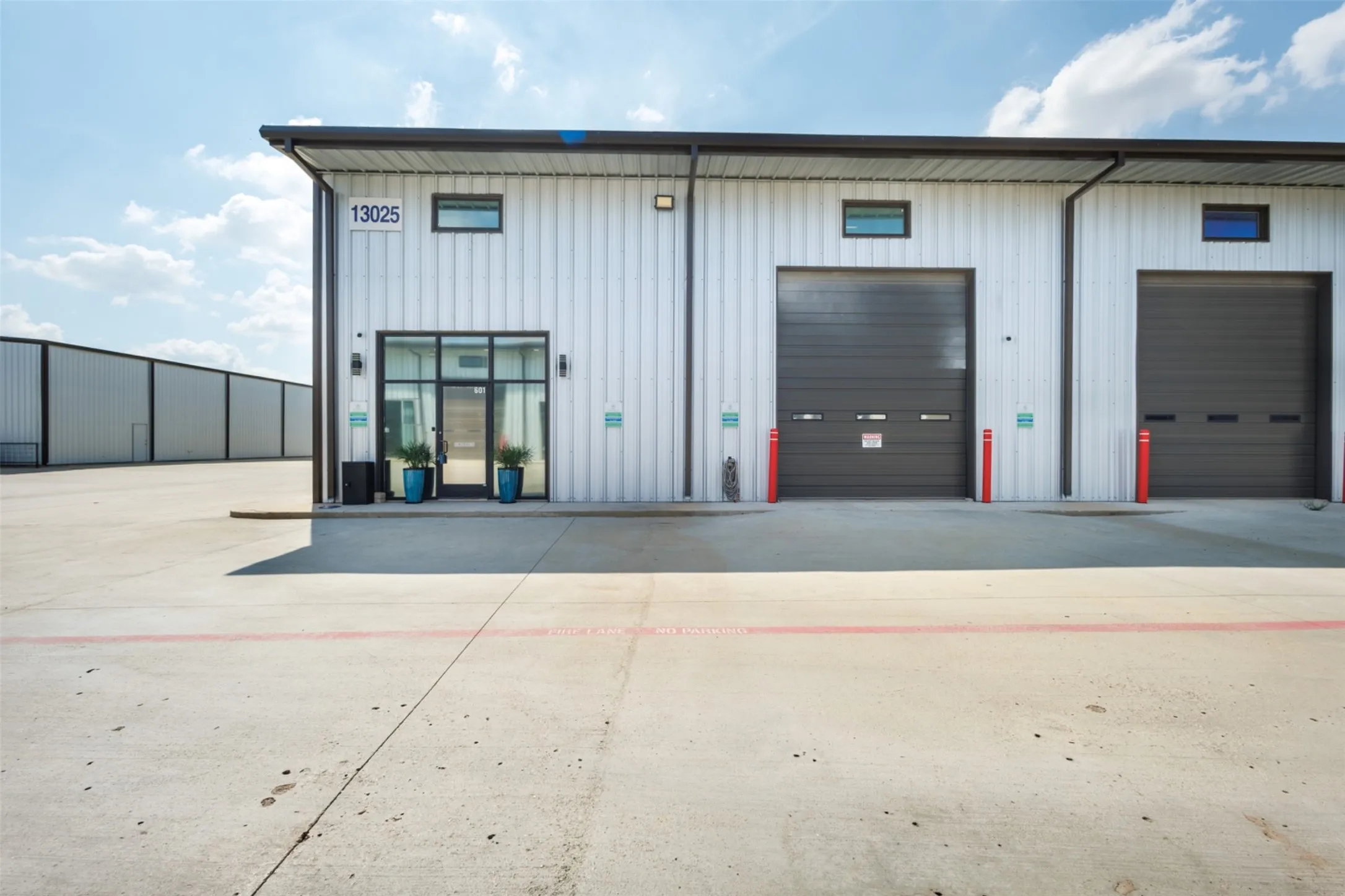 Corrugated metal exterior with multiple roll-up doors, large concrete paved area, and modern glass entry