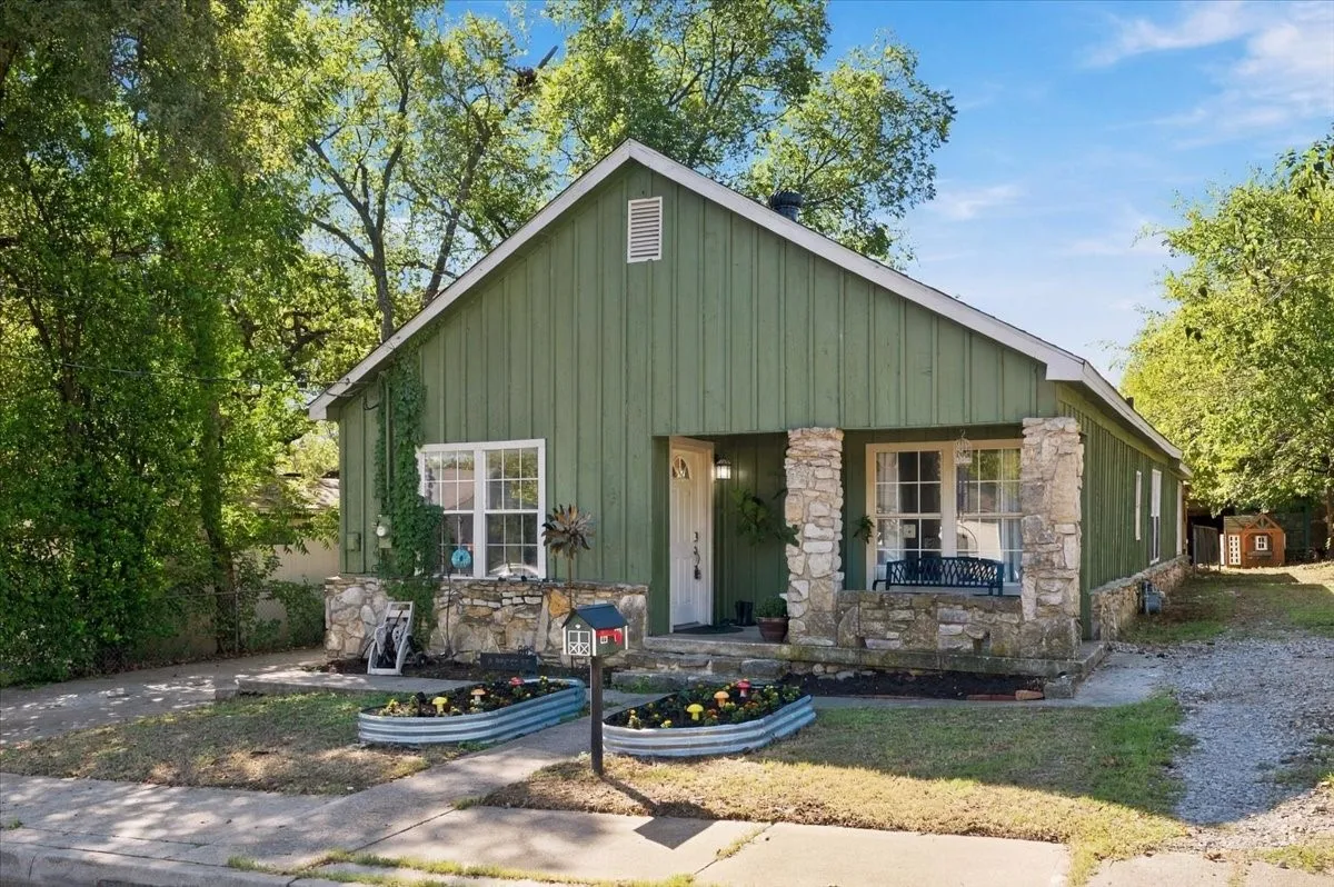 Modern farmhouse residence featuring a distinctive green metal exterior with board-and-batten siding, stone chimney accents, and a covered front porch with stone pillars