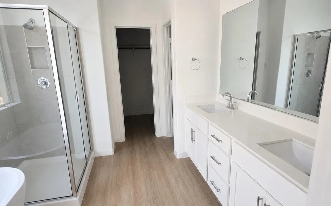 Bathroom featuring a dual vanity with white cabinetry and integrated sinks, a glass-enclosed shower with tiled walls and a built-in niche, and wood-finish flooring