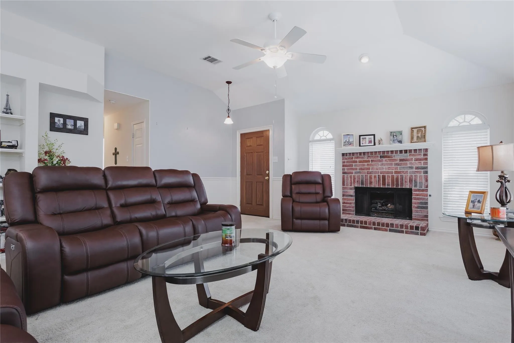 Vaulted ceilings, brick fireplace, built-in shelving, arched windows with blinds, and a ceiling fan with integrated lighting