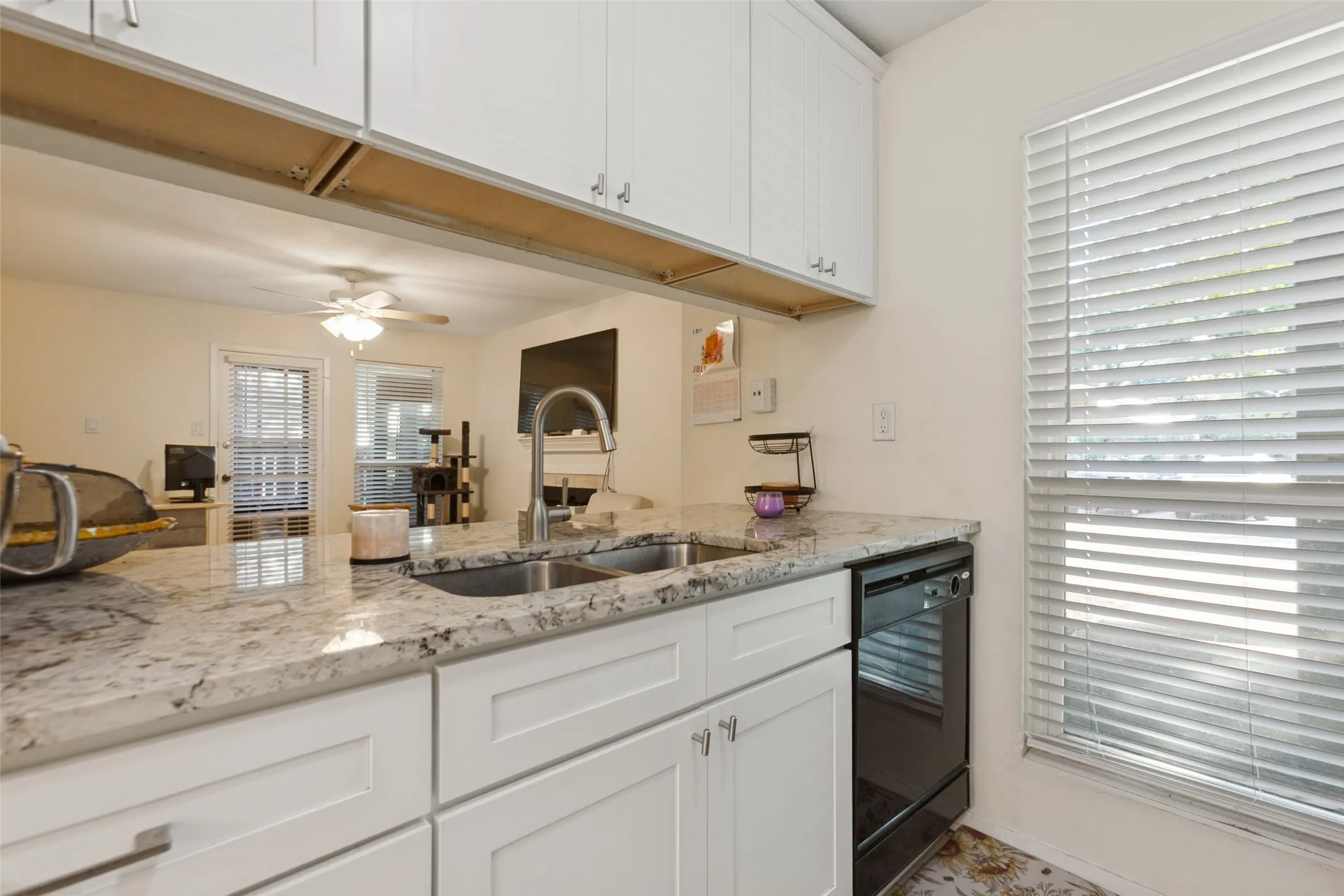 White shaker-style cabinetry with chrome hardware, dual basin stainless steel sink with a gooseneck faucet, light-toned countertop, and a black dishwasher