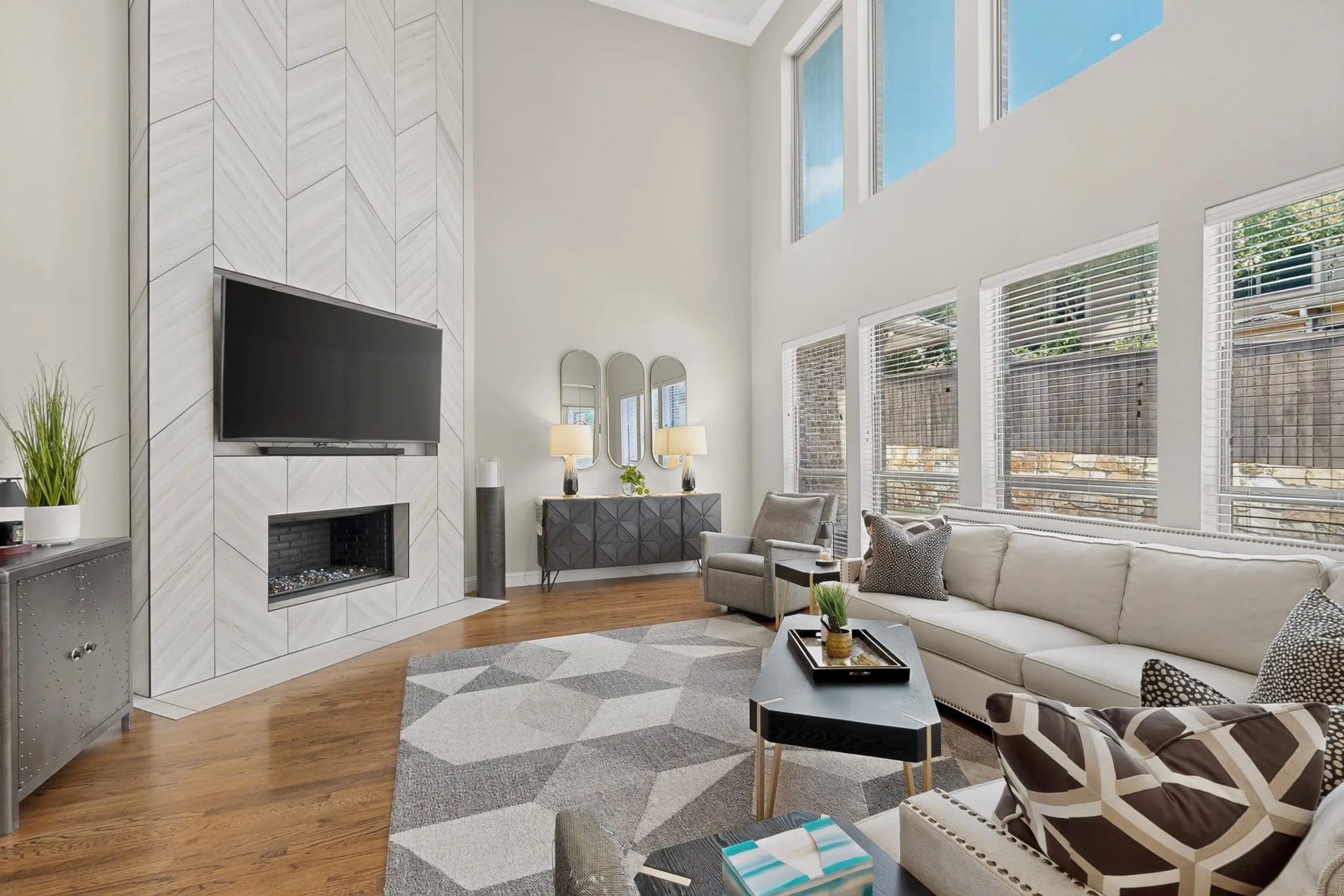 Two-story living room featuring a herringbone-patterned fireplace with an integrated linear firebox, natural wood-finish flooring, and expansive windows