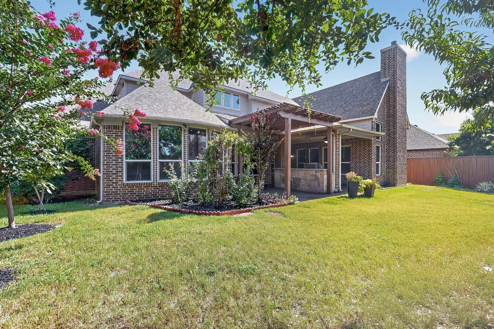 Expansive backyard featuring a manicured lawn, a covered patio with a built-in outdoor kitchen, an attached pergola, a brick chimney, and a sunroom with multiple windows