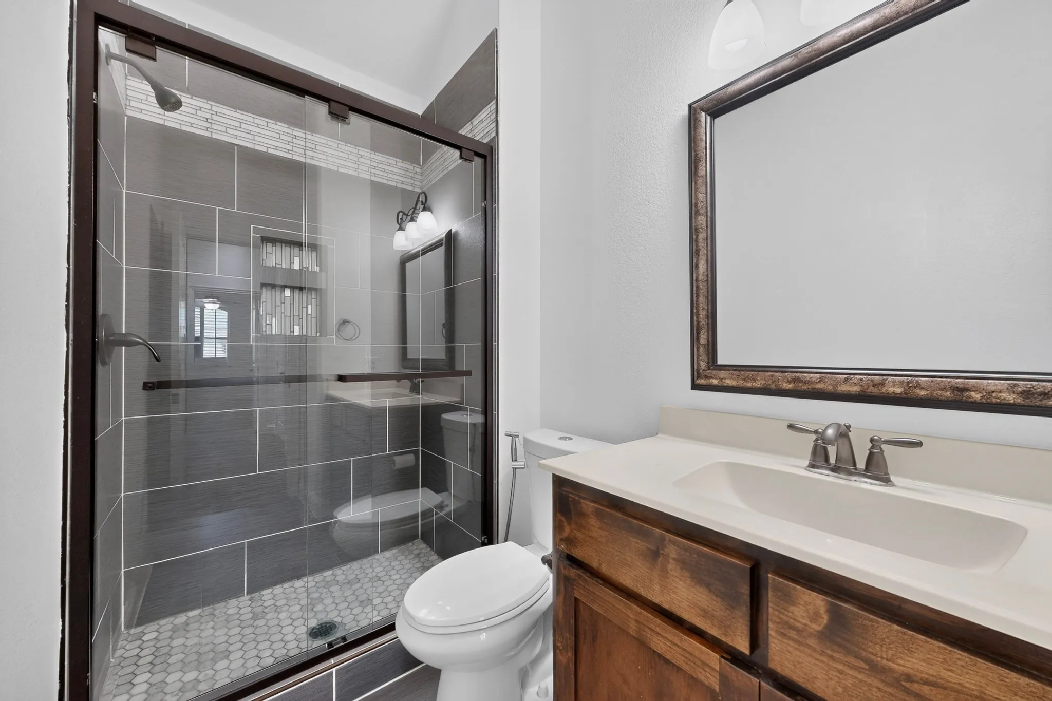 Glass-enclosed shower with dark tile surround and mosaic floor, wood-finish vanity with an integrated sink, framed mirror, and brushed nickel fixtures