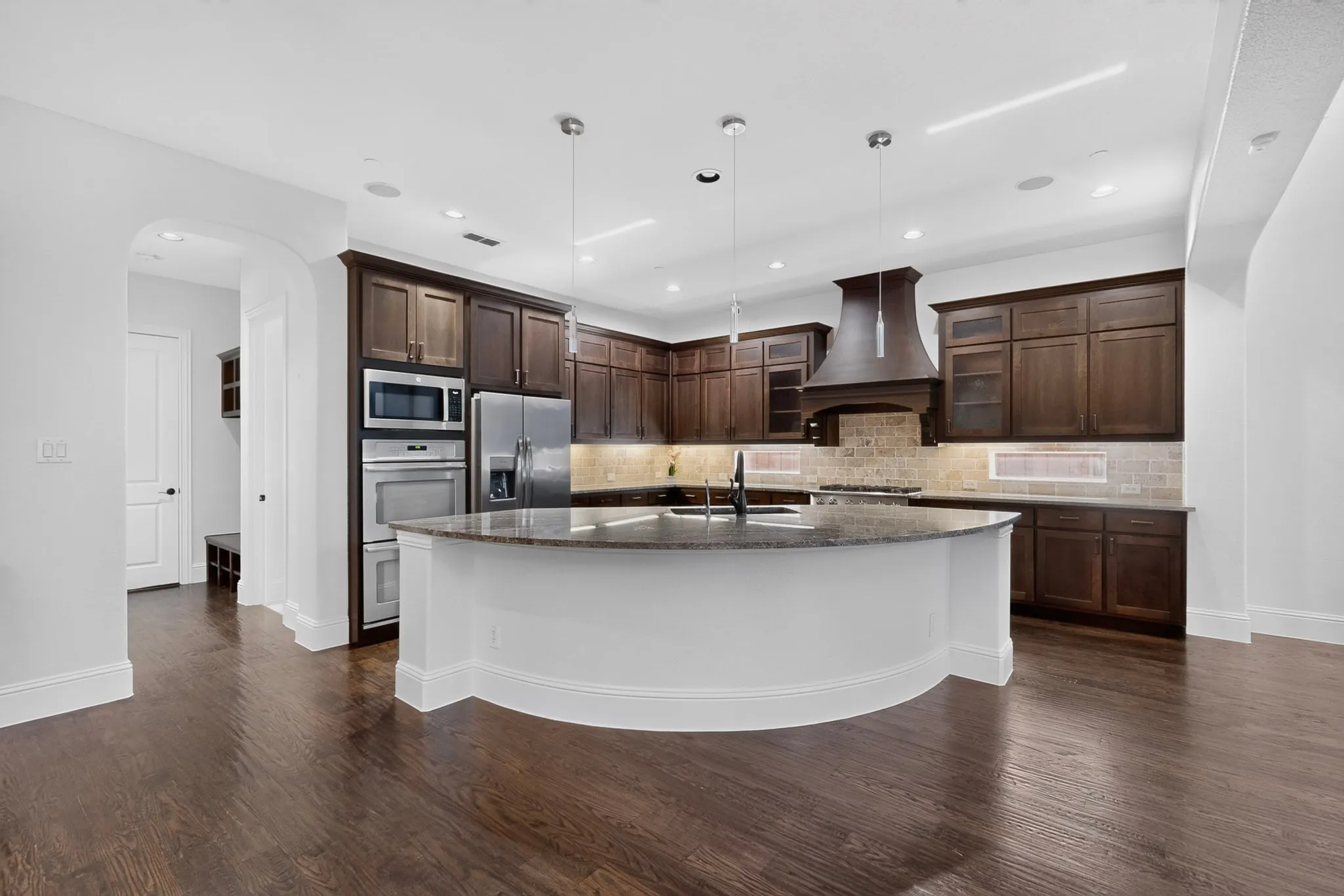 Spacious kitchen featuring an expansive curved island with a stone countertop, dark wood-finish cabinetry, stainless steel appliances, and wood-finish flooring