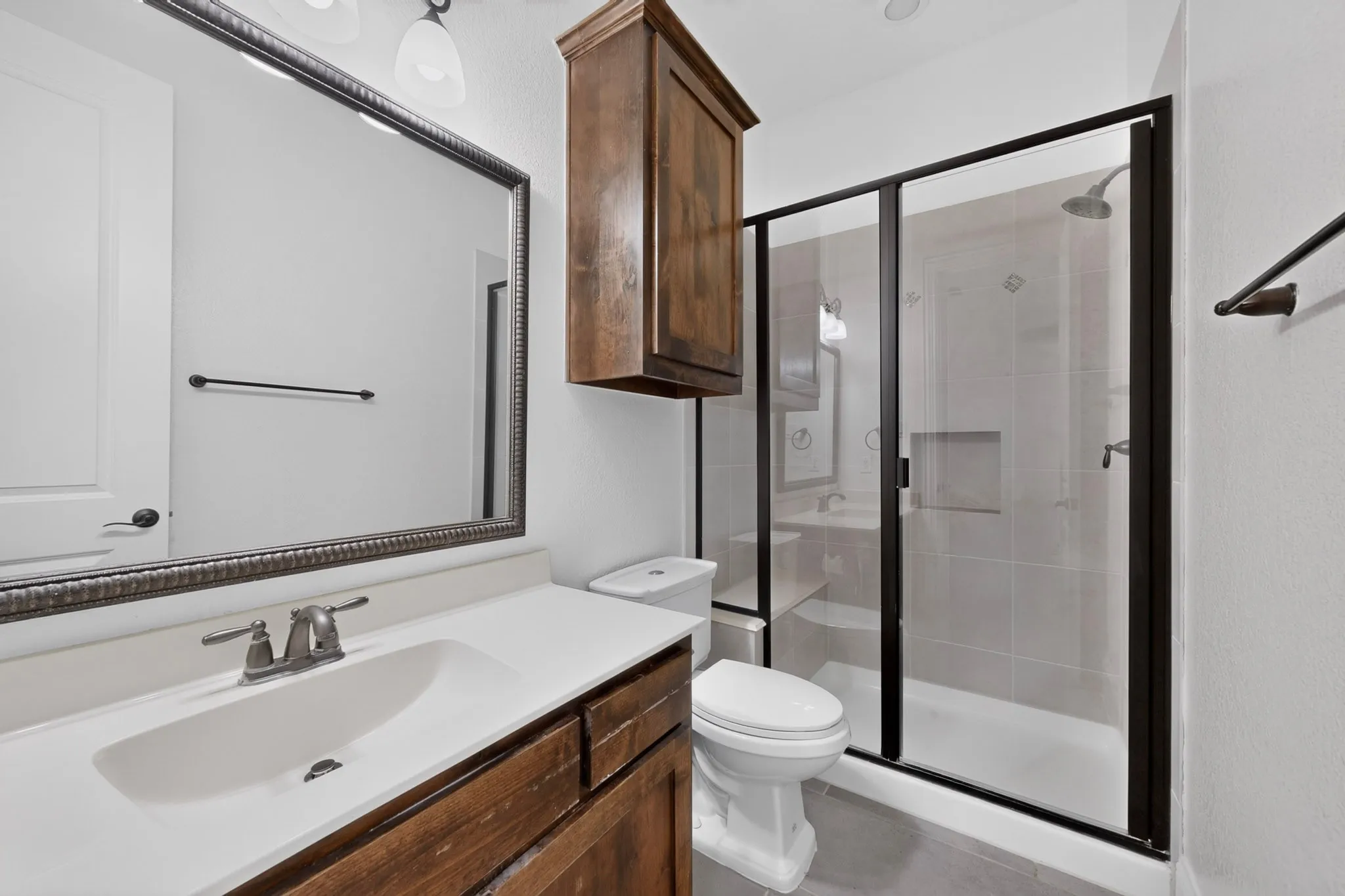 Bathroom featuring a spacious walk-in shower with clear glass enclosure and built-in niche, a single vanity with an integrated sink and brushed nickel faucet, a large framed mirror, a wall-mounted wooden storage cabinet, and white walls