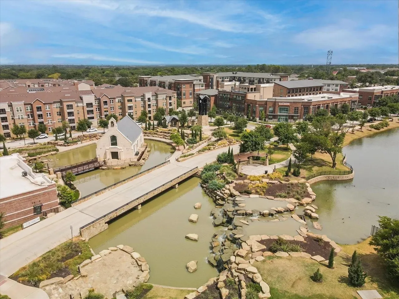 Waterfront mixed-use development featuring brick and stone architectural elements, manicured landscaping with mature trees, and direct water access