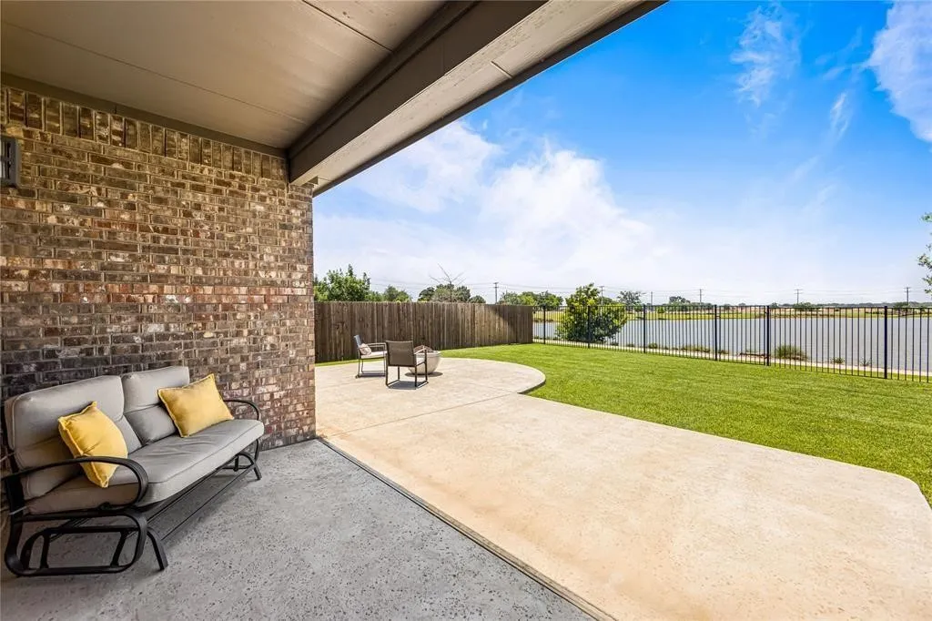 Covered patio with brick wall, expansive concrete patio, manicured lawn, and black metal fencing along water