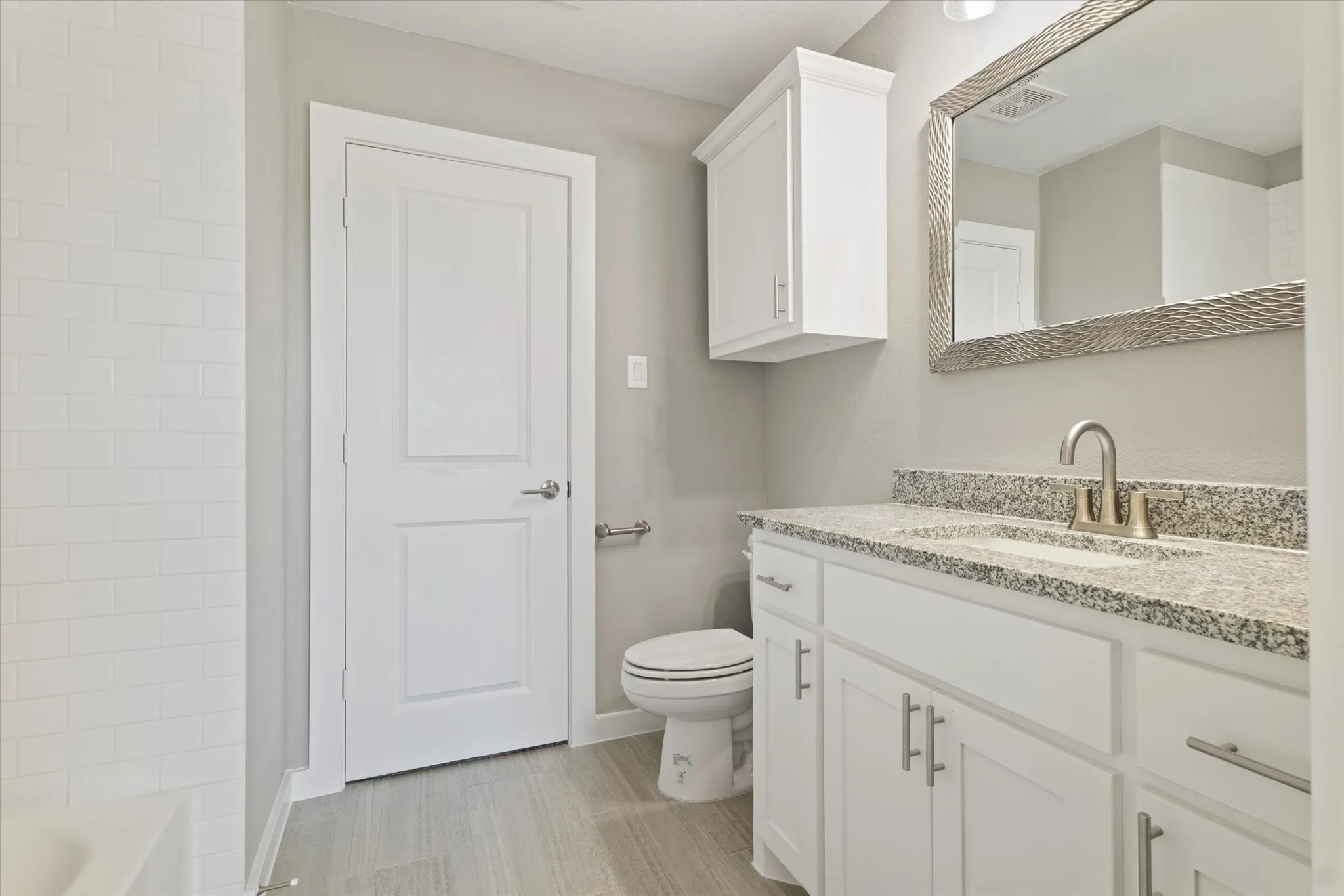 White subway tile surround, wood-finish flooring, white vanity with stone countertop, stainless steel sink with single-handle faucet, and mirrored wall cabinet