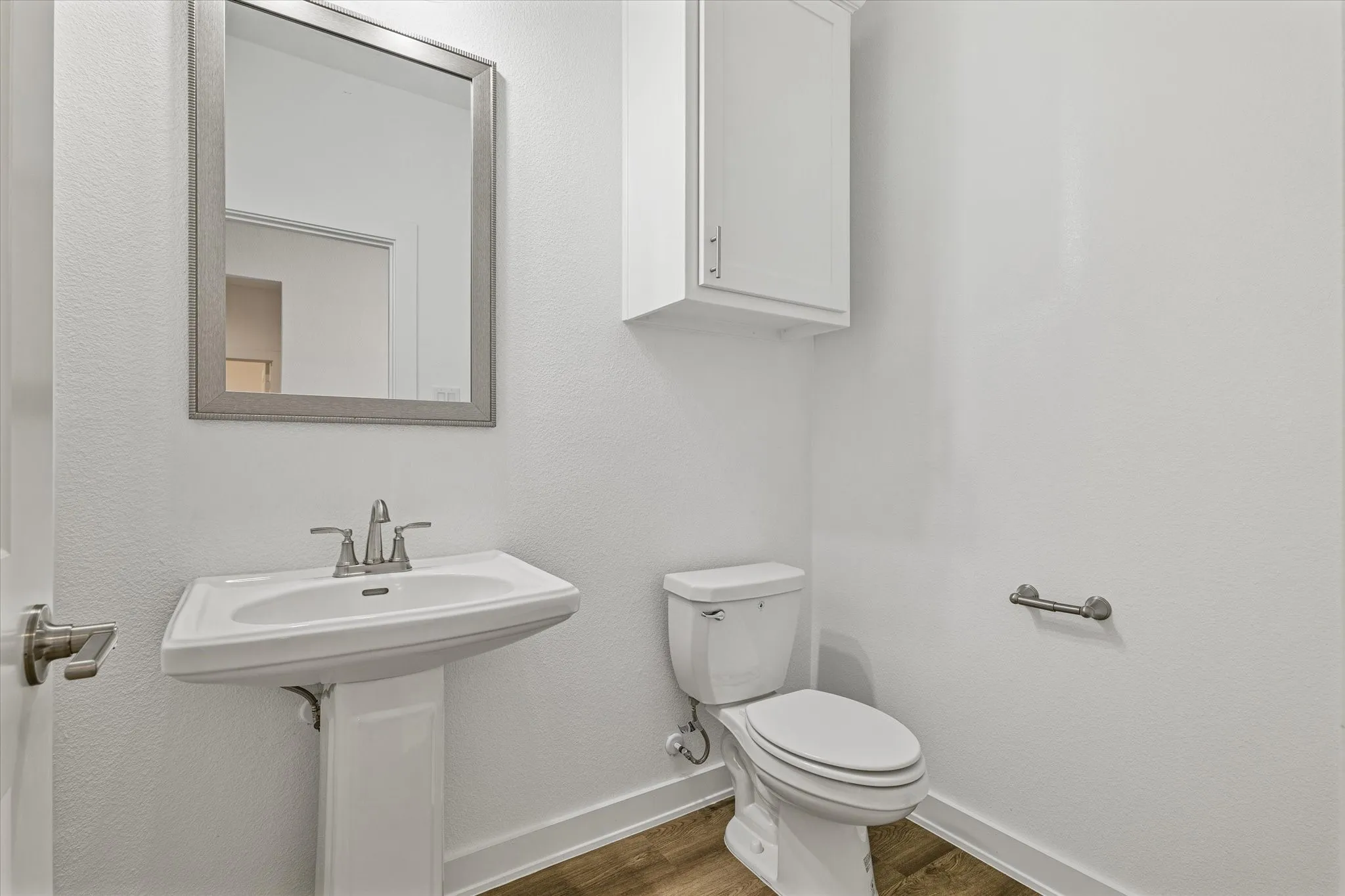Pedestal sink with a chrome faucet, wall-mounted mirror, white toilet, and wood-finish flooring