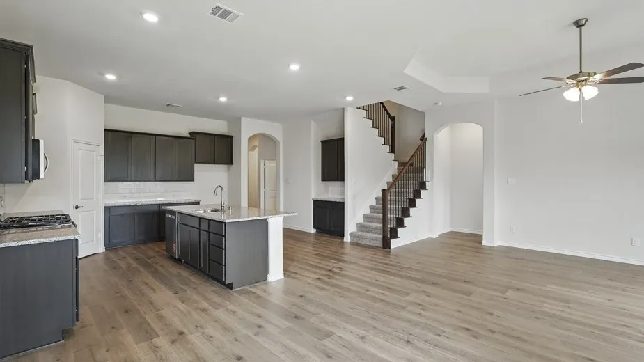 Open-concept living space featuring wood-finish flooring, a contemporary kitchen with dark cabinetry and a central island, a multi-level ceiling with a ceiling fan, a carpeted staircase with an iron spindle railing, and recessed lighting
