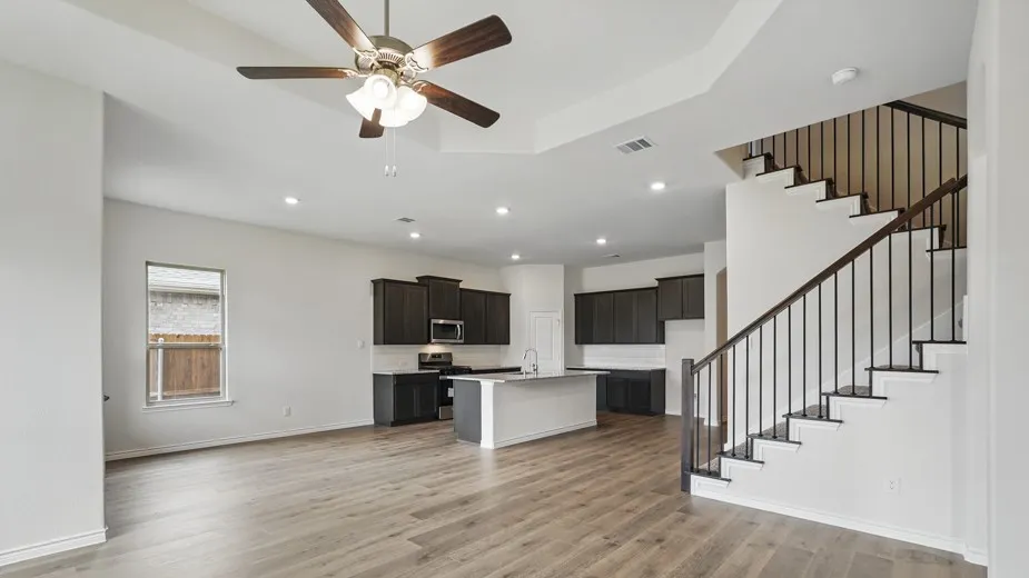Open-concept interior featuring wood-finish flooring, a contemporary kitchen with dark cabinetry and a white island, a staircase with dark wood handrails and black metal balusters, recessed lighting, and a ceiling fan