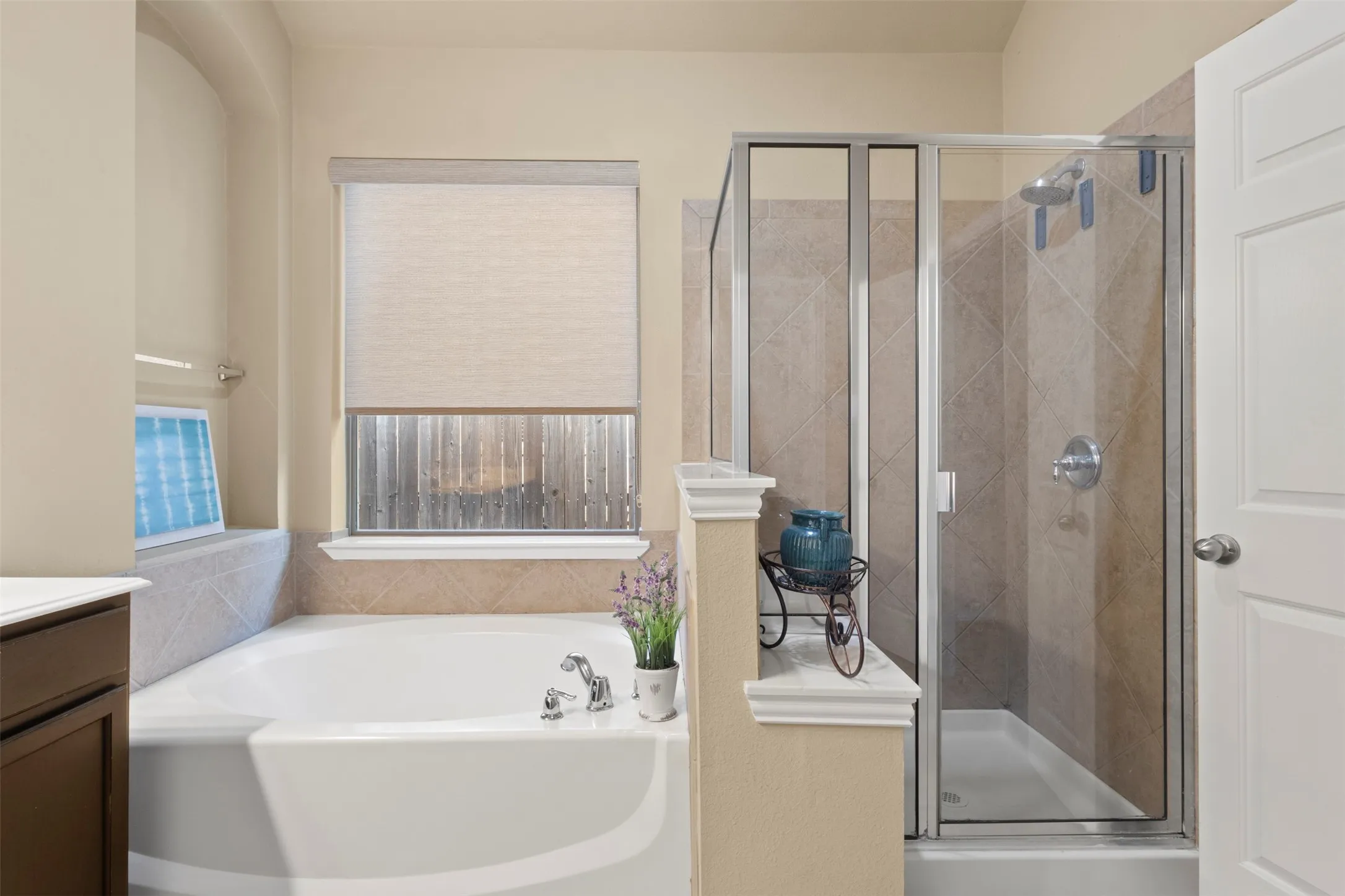 Built-in bathtub, glass-enclosed shower, tiled shower surround, window with roller shade, and built-in vanity cabinetry