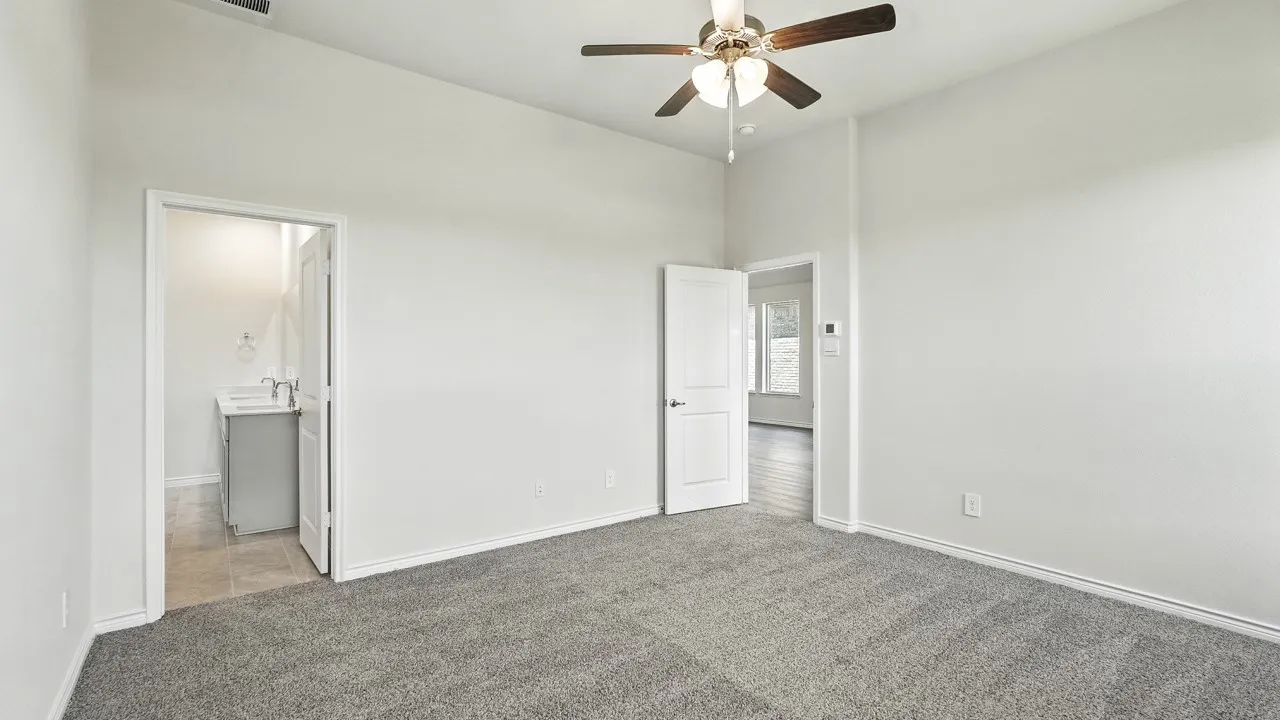 Spacious room featuring neutral carpeting, light gray walls, a ceiling fan with integrated lighting, and a white paneled door accessing a bathroom with a modern vanity