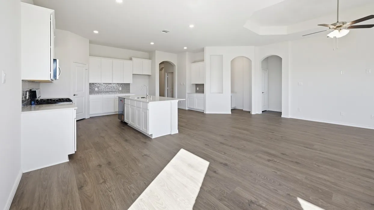 Open-concept interior featuring light wood-finish flooring, a contemporary kitchen with white cabinetry and stainless steel appliances, a center island with sink, a decorative tile backsplash, and a spacious living area with a tray ceiling and ceiling fan