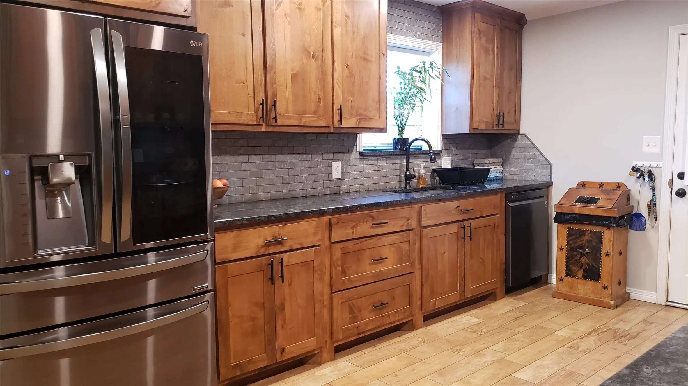 Stainless steel refrigerator, wood-finish cabinetry, dark countertops, tiled backsplash, and wood-finish flooring