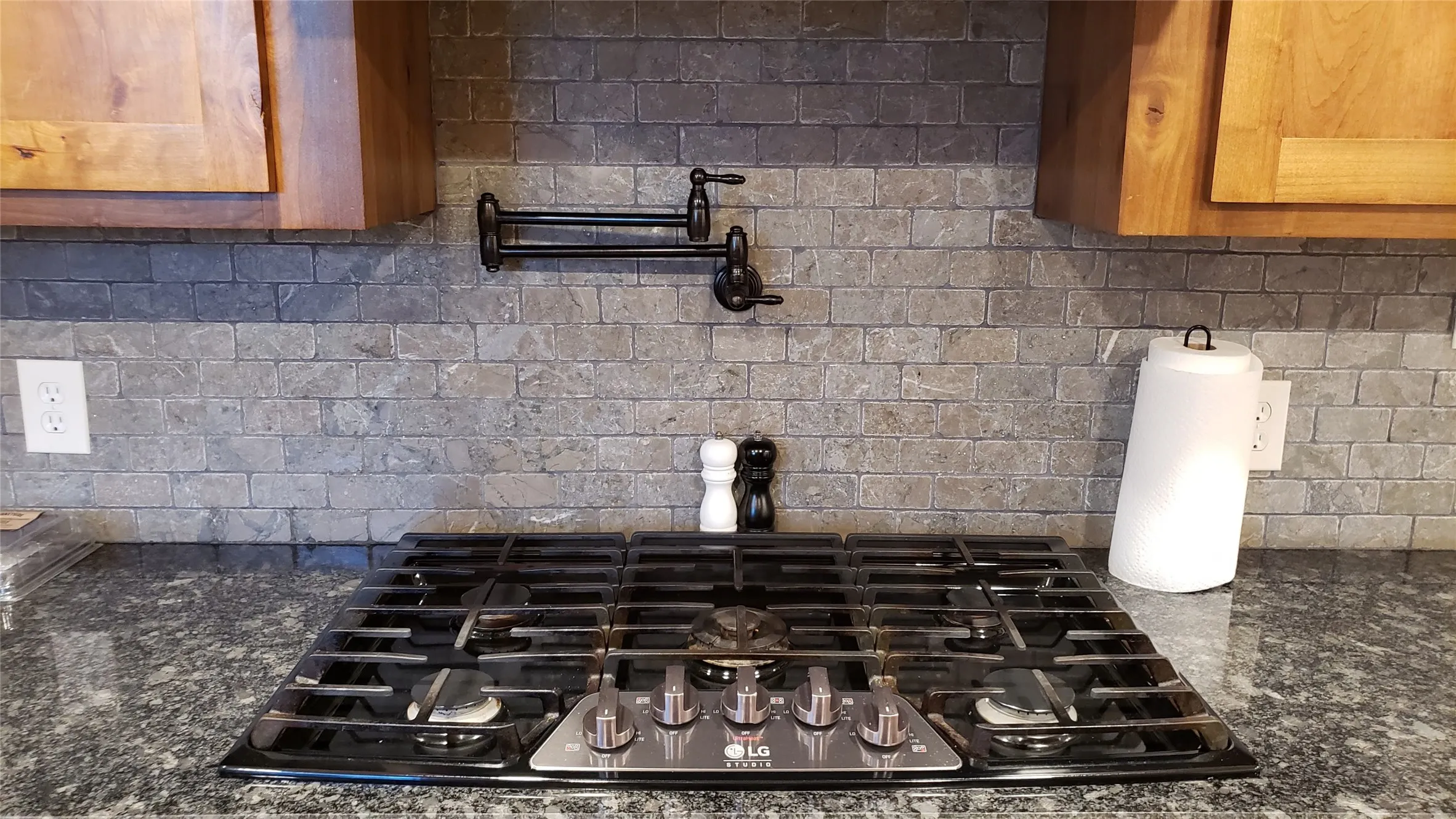 Gas cooktop, stone-finish backsplash, dark granite-style countertops, wood cabinetry, and a wall-mounted pot filler