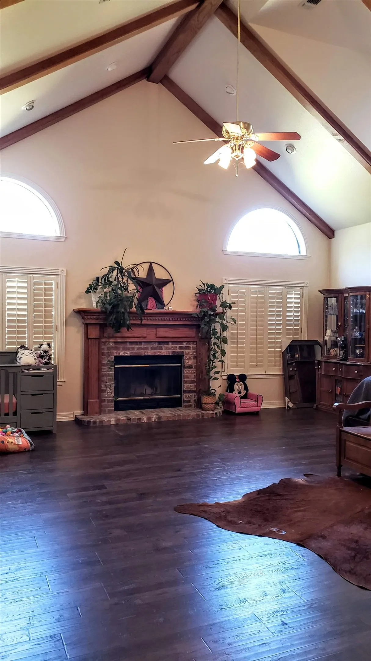 Vaulted ceiling with exposed wood beams, brick fireplace with wood mantel, arched transom windows, and wood-finish flooring