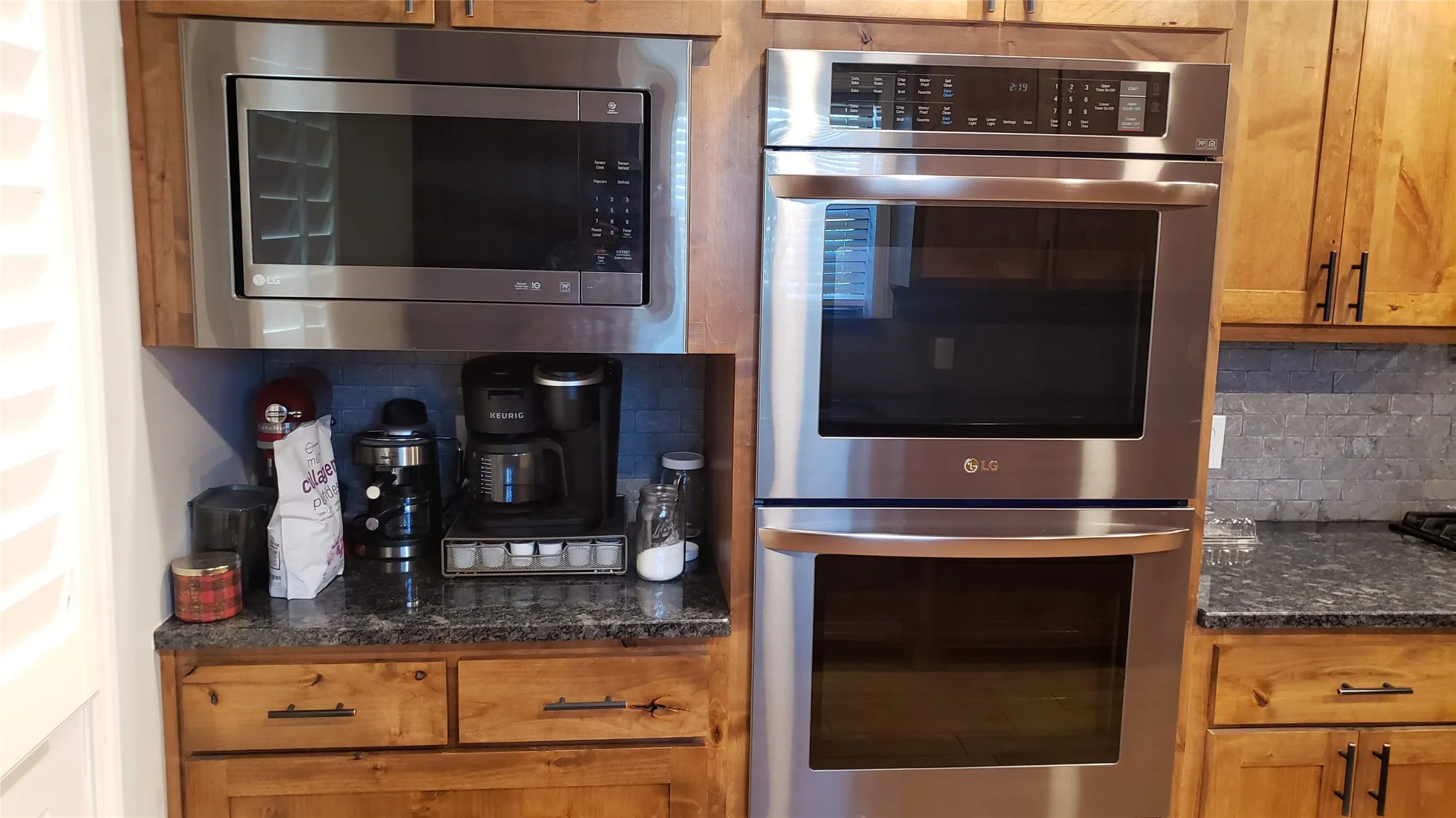 Stainless steel microwave, double wall oven, wood cabinetry, stone-finish countertop, and tile backsplash