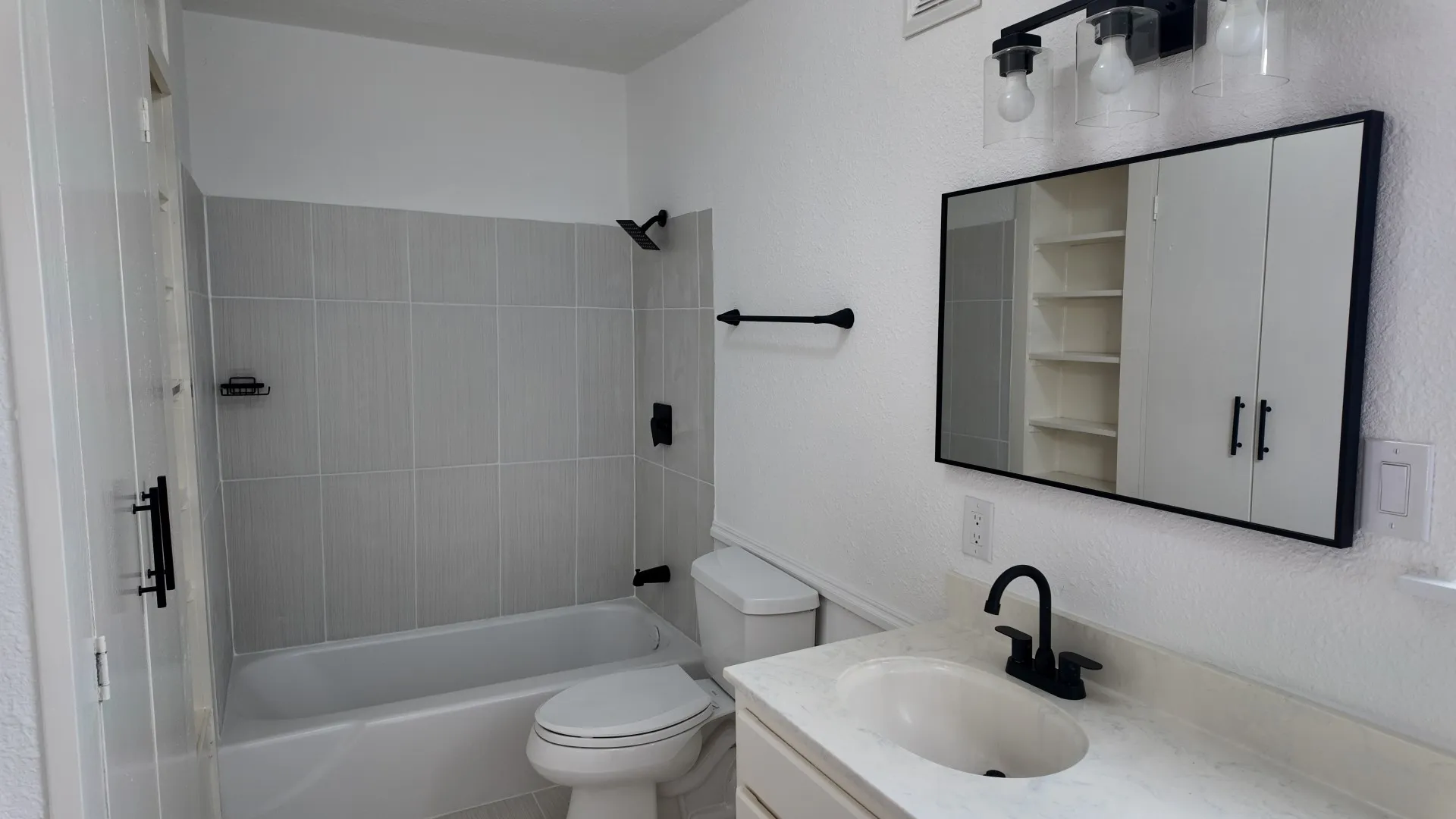 White bathtub with light gray vertical tile surround, matte black shower fixtures, vanity with integrated sink and matte black faucet, and a wall-mounted mirrored cabinet