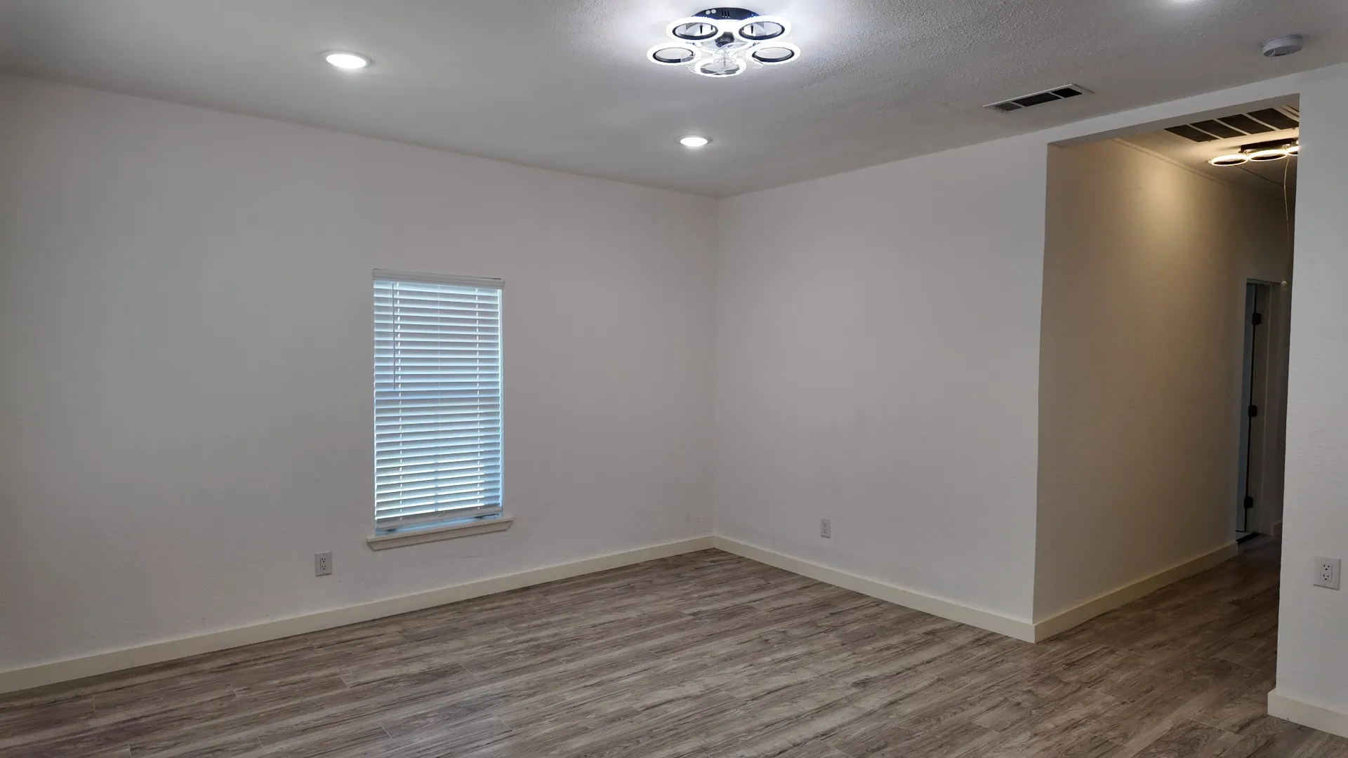 Recessed lighting and modern ceiling fixtures illuminate the room with wood-finish flooring and a single window with blinds