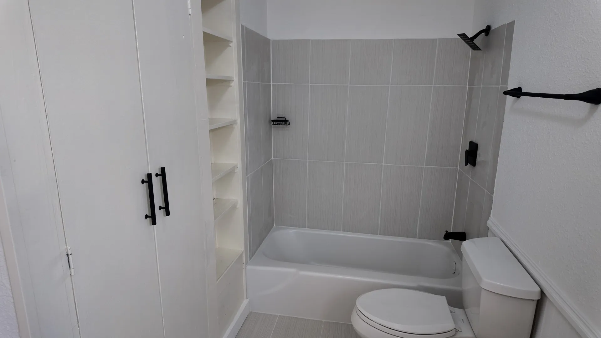 White built-in cabinetry with open shelving, a white bathtub, a toilet, light gray subway tile, and matte black fixtures